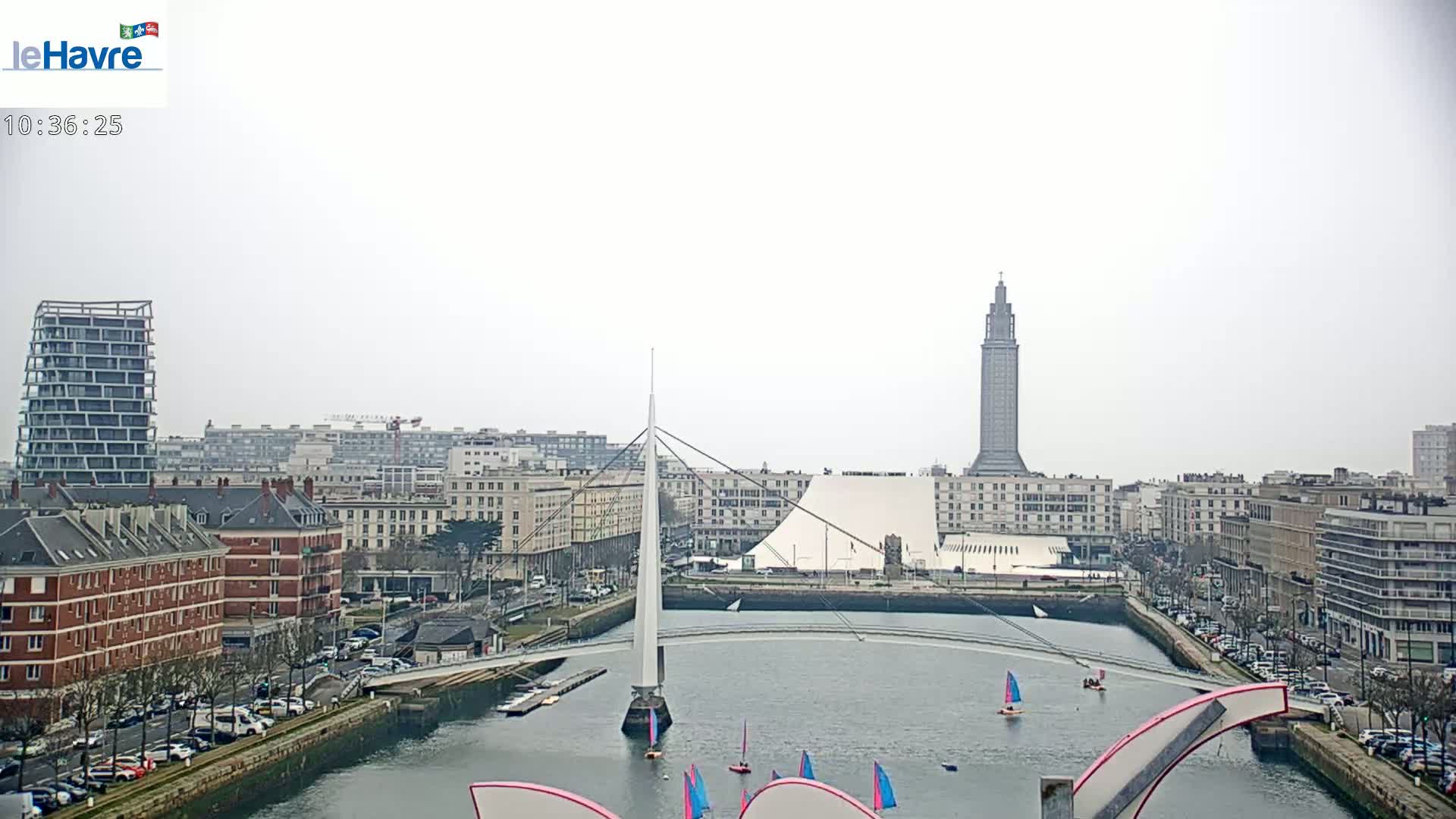 A city waterfront on an overcast day features a tall tower, a modern white building, a cable-stayed bridge, and sailboats on the water.