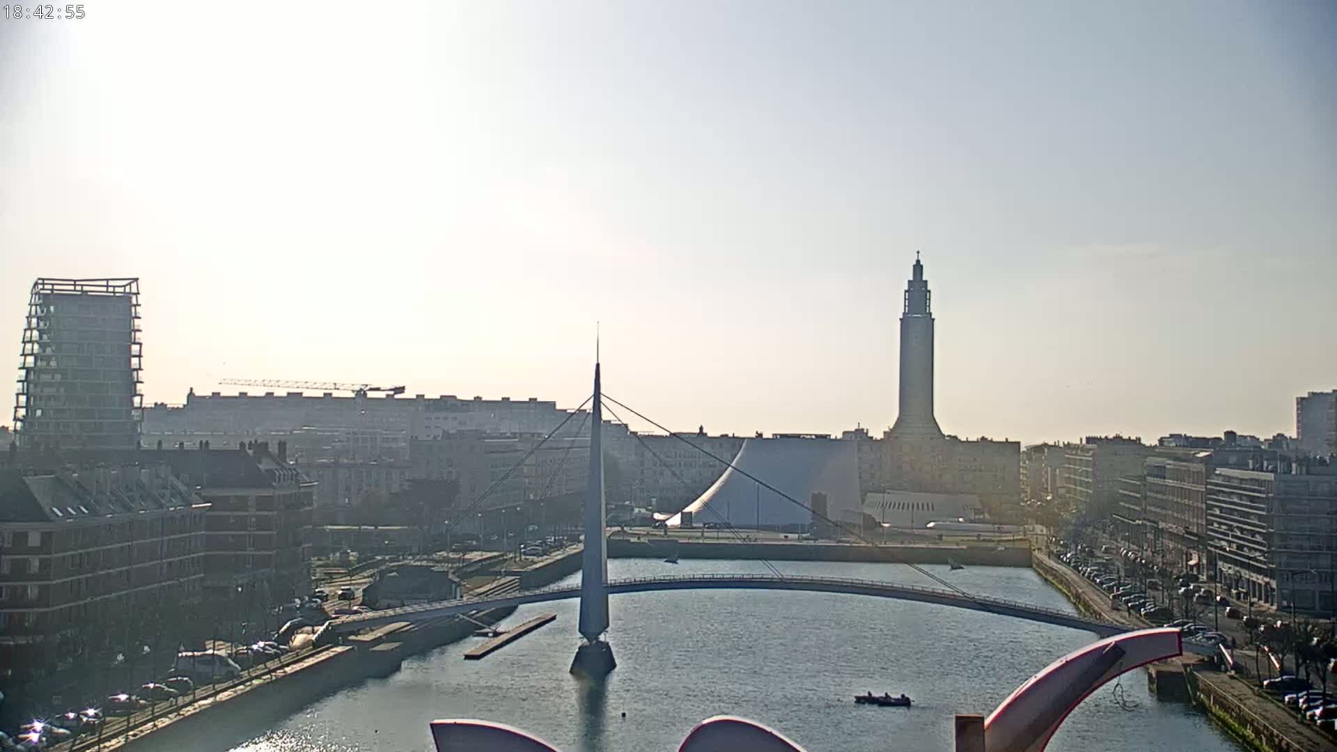 A city waterfront scene on a bright, sunny day shows a canal with a modern pedestrian bridge, buildings of varying heights, and a tall tower in the background.