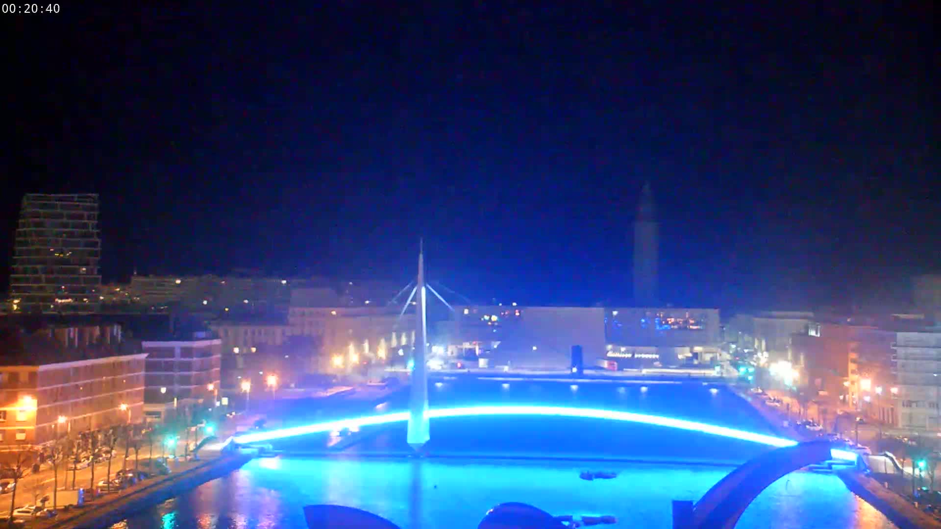 A nighttime view of a city waterfront with a modern bridge illuminated in blue, reflecting in the calm water.