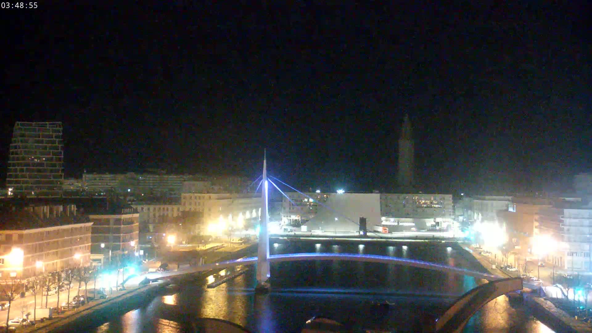 A nighttime view of a city with a modern, illuminated bridge spanning a waterway, under a clear, dark sky.