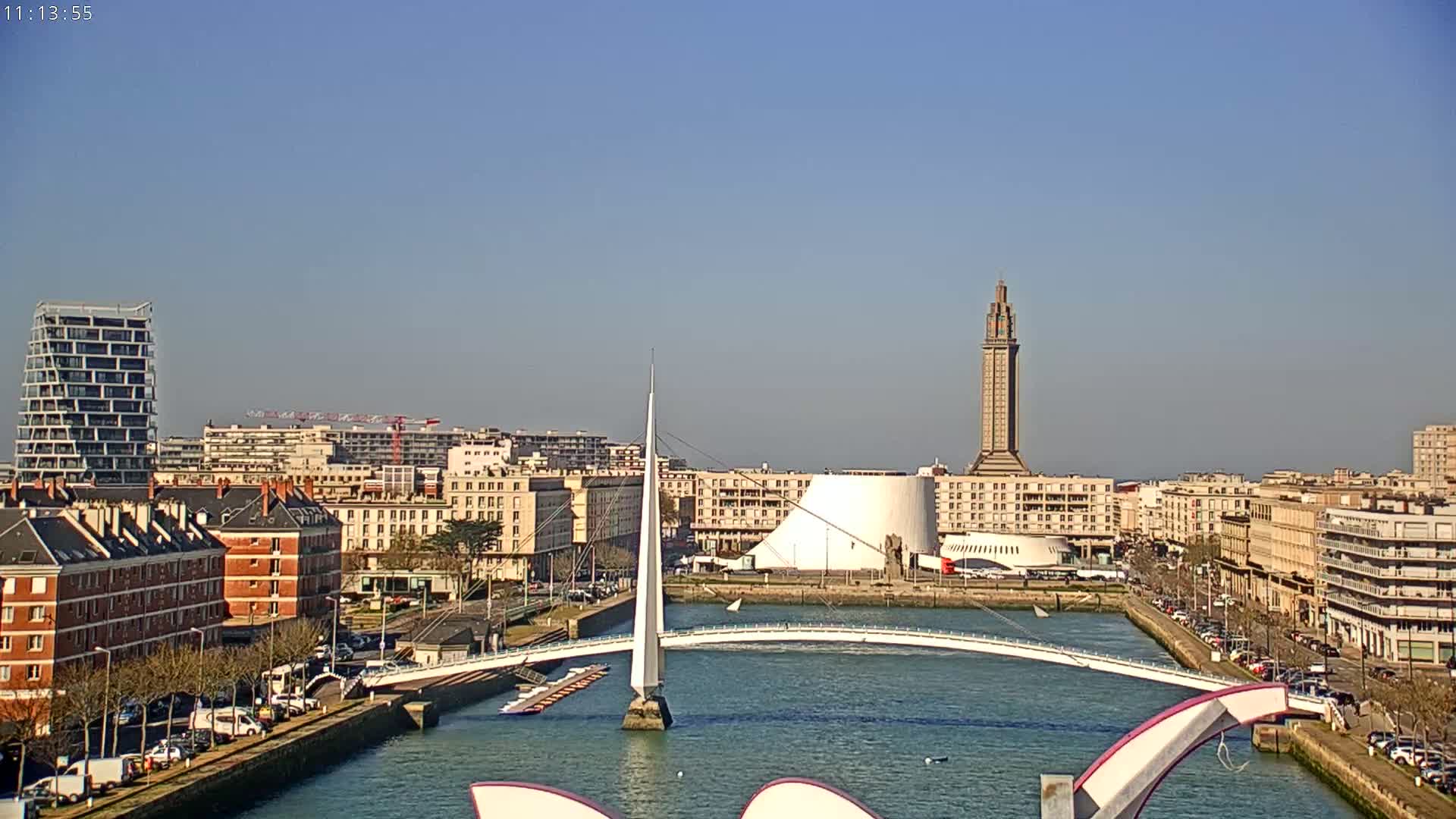 A cityscape with a modern white bridge spanning a waterway, under a clear blue sky, features a tall tower in the mid-ground and various buildings along the waterfront.