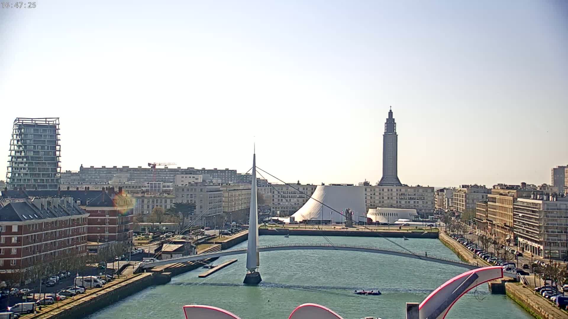 A city waterfront scene on a sunny day shows a canal with a modern pedestrian bridge, buildings, and a tall tower in the background.