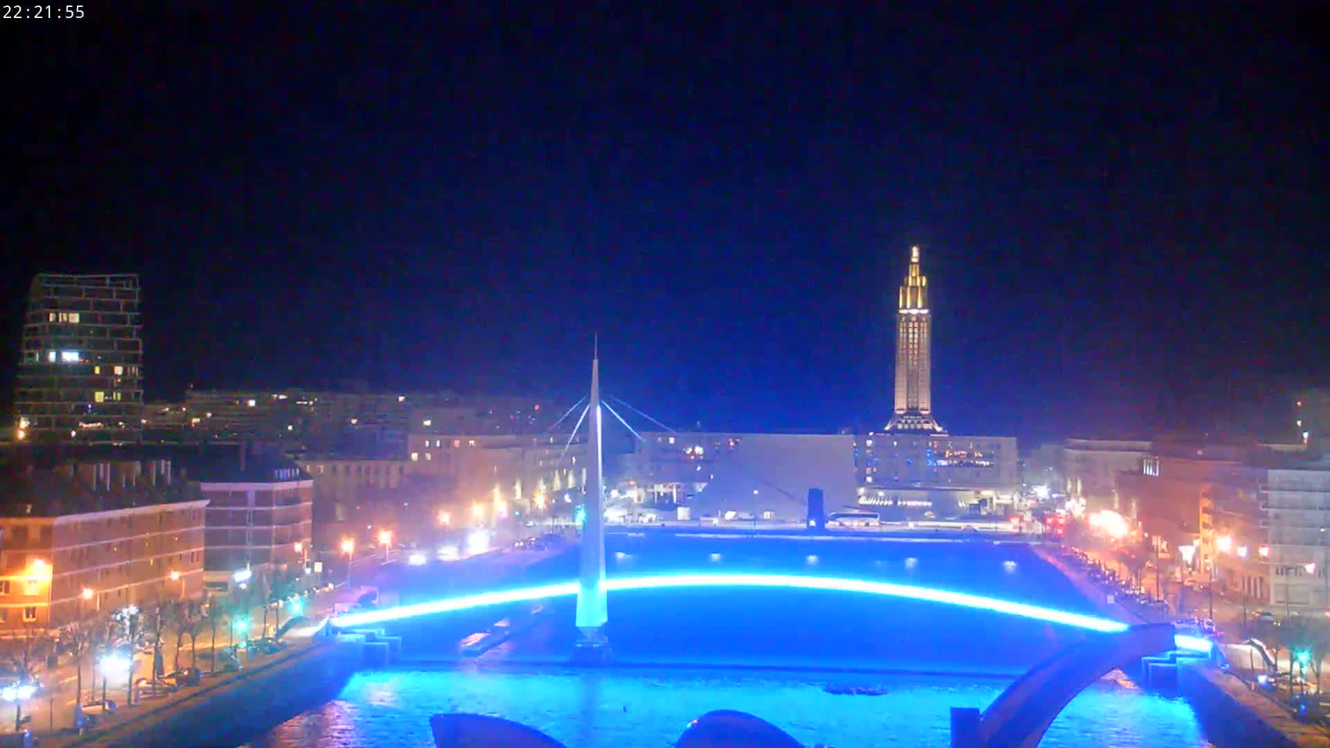 A city at night, illuminated with lights, features a tall tower in the distance and a bridge with blue lights spanning a body of water.