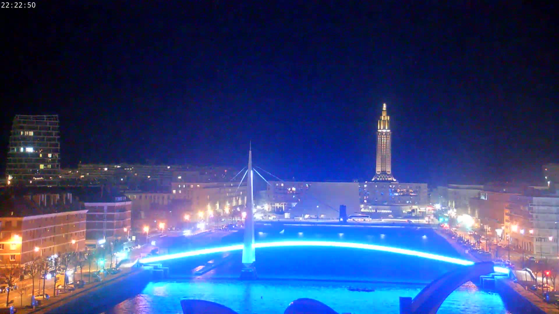 A nighttime cityscape features a bridge illuminated in blue light over a body of water, with city buildings and a tall tower visible in the background under a clear, dark sky.