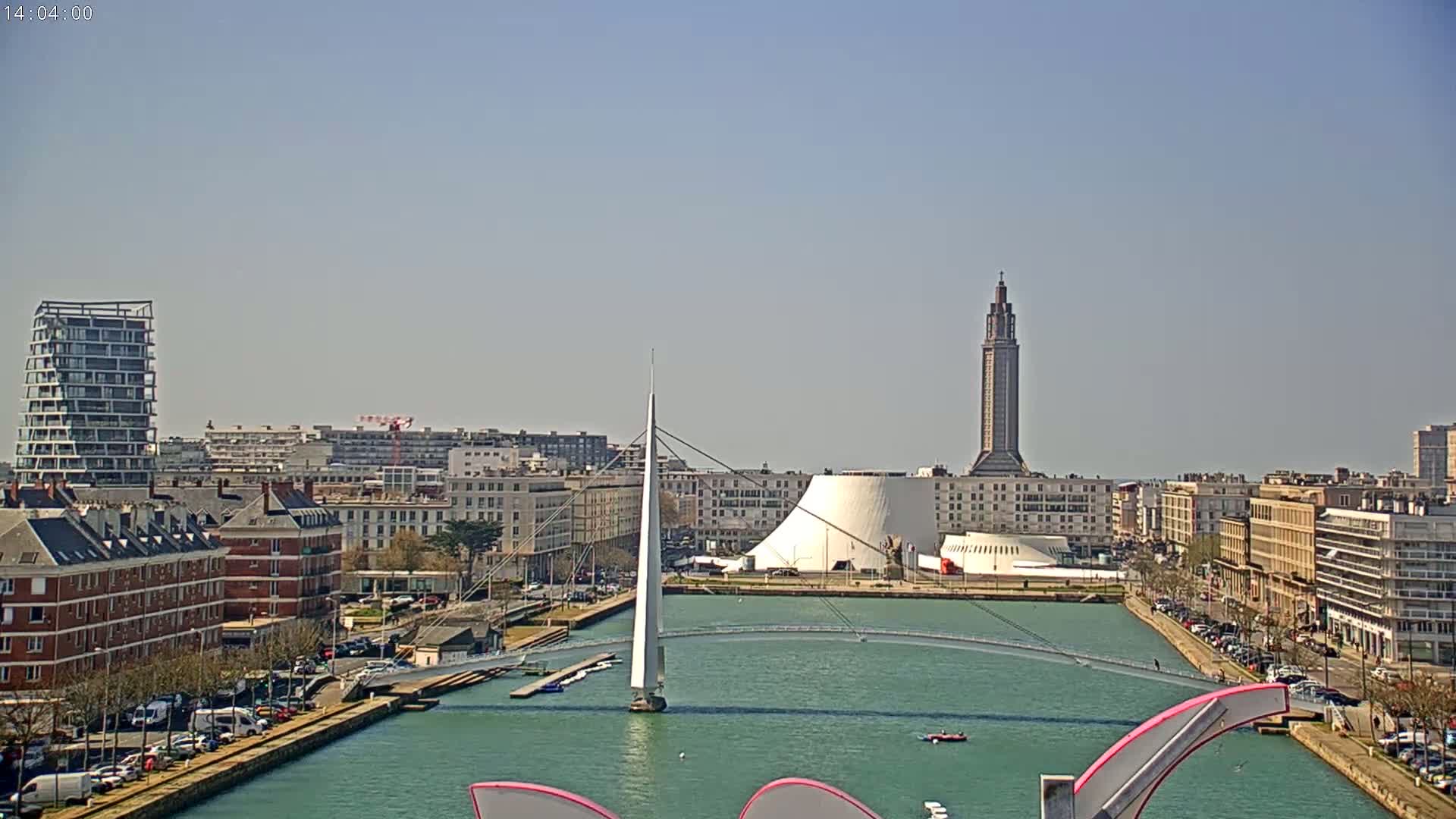 A waterfront city scene on a sunny day shows a canal with a modern white pedestrian bridge, buildings of varying heights and styles, and a tall tower in the background.