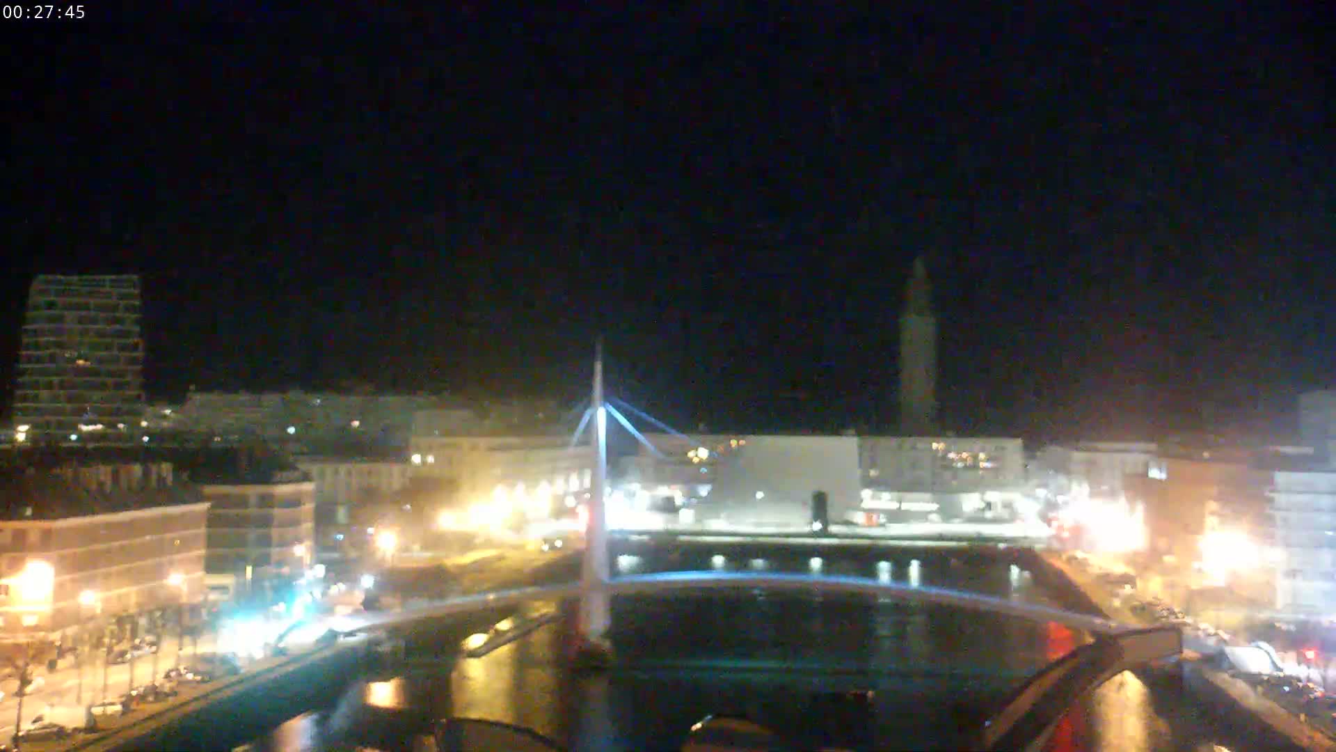 A nighttime view of a city with a bridge illuminated over a waterway, under a clear, dark sky.