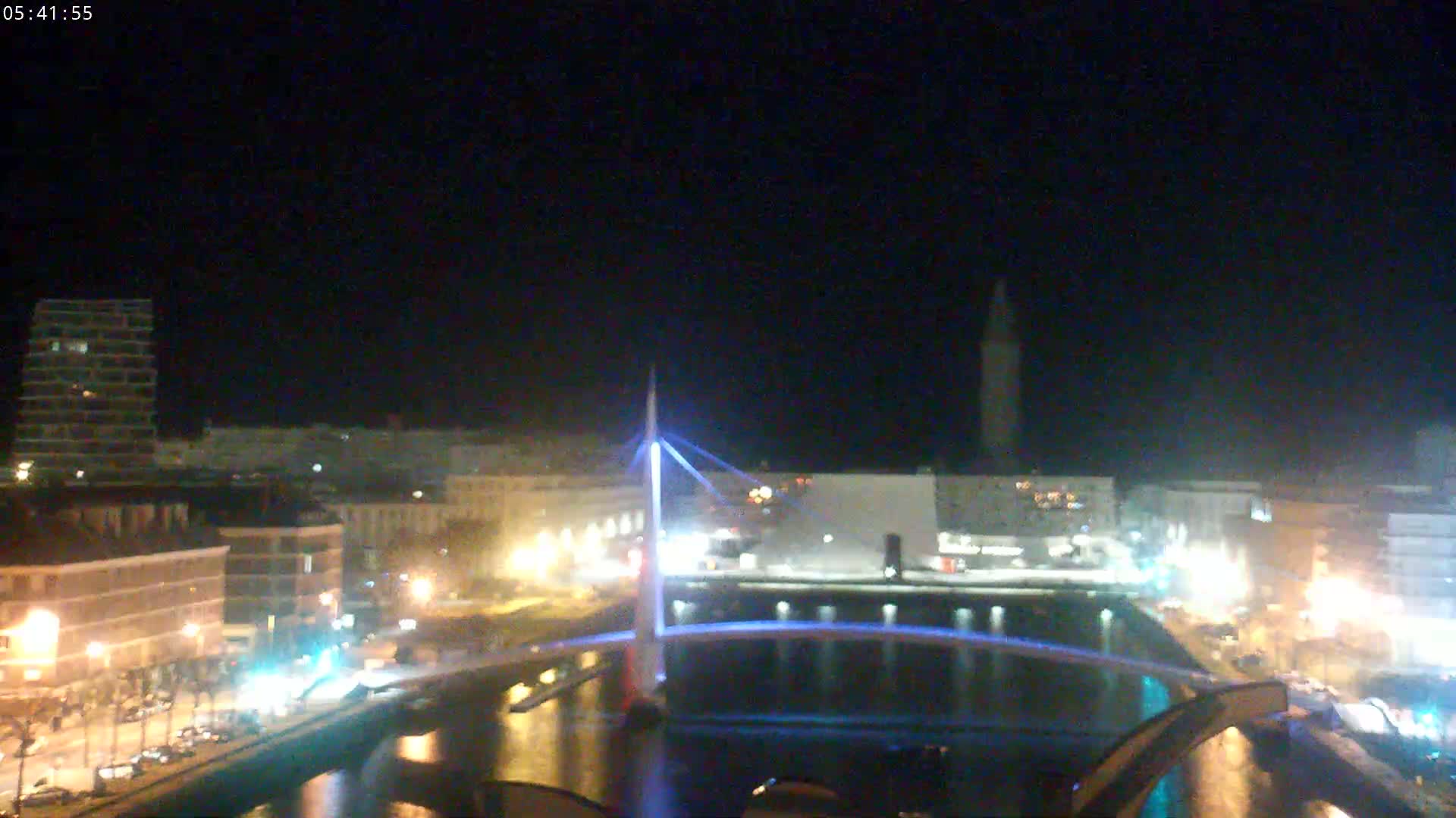 A nighttime view of a city's illuminated waterfront, featuring a bridge with blue lights and buildings lining a canal.