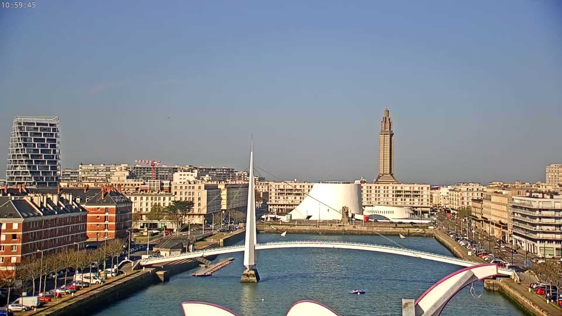 A city waterfront on a sunny day shows a white pedestrian bridge arching over a waterway, with buildings, a tall tower, and parked cars lining the shore.