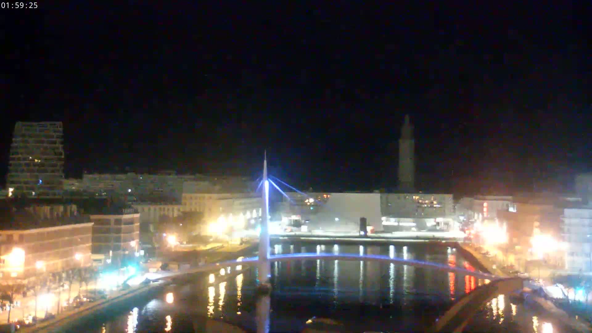 A nighttime view of a city waterfront showing a lit bridge spanning a calm canal, buildings along the shoreline, and a tall structure in the distance under a clear, dark sky.