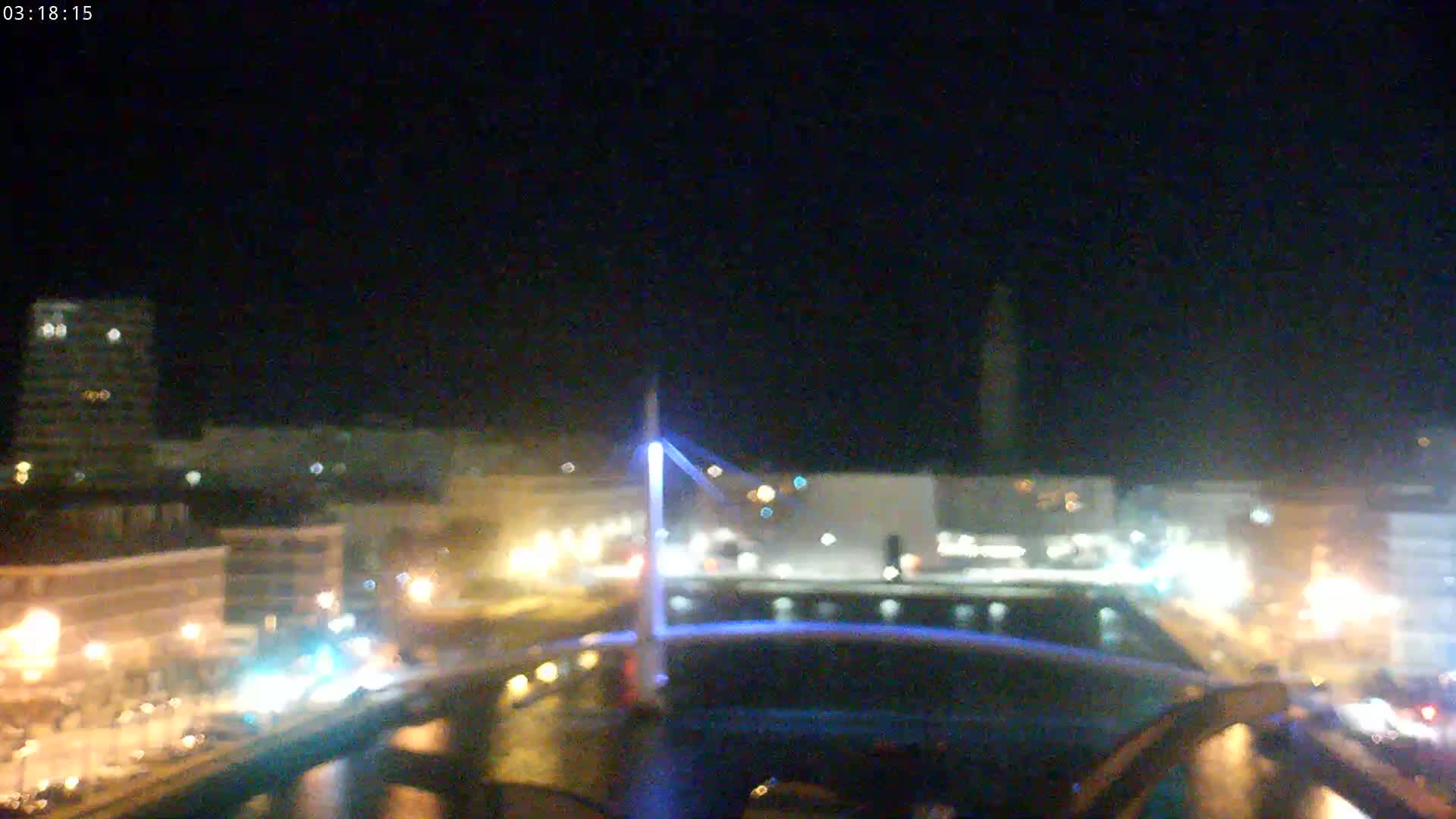 A nighttime view of a city waterfront with a bridge illuminated in blue, showing city lights reflecting on dark water.