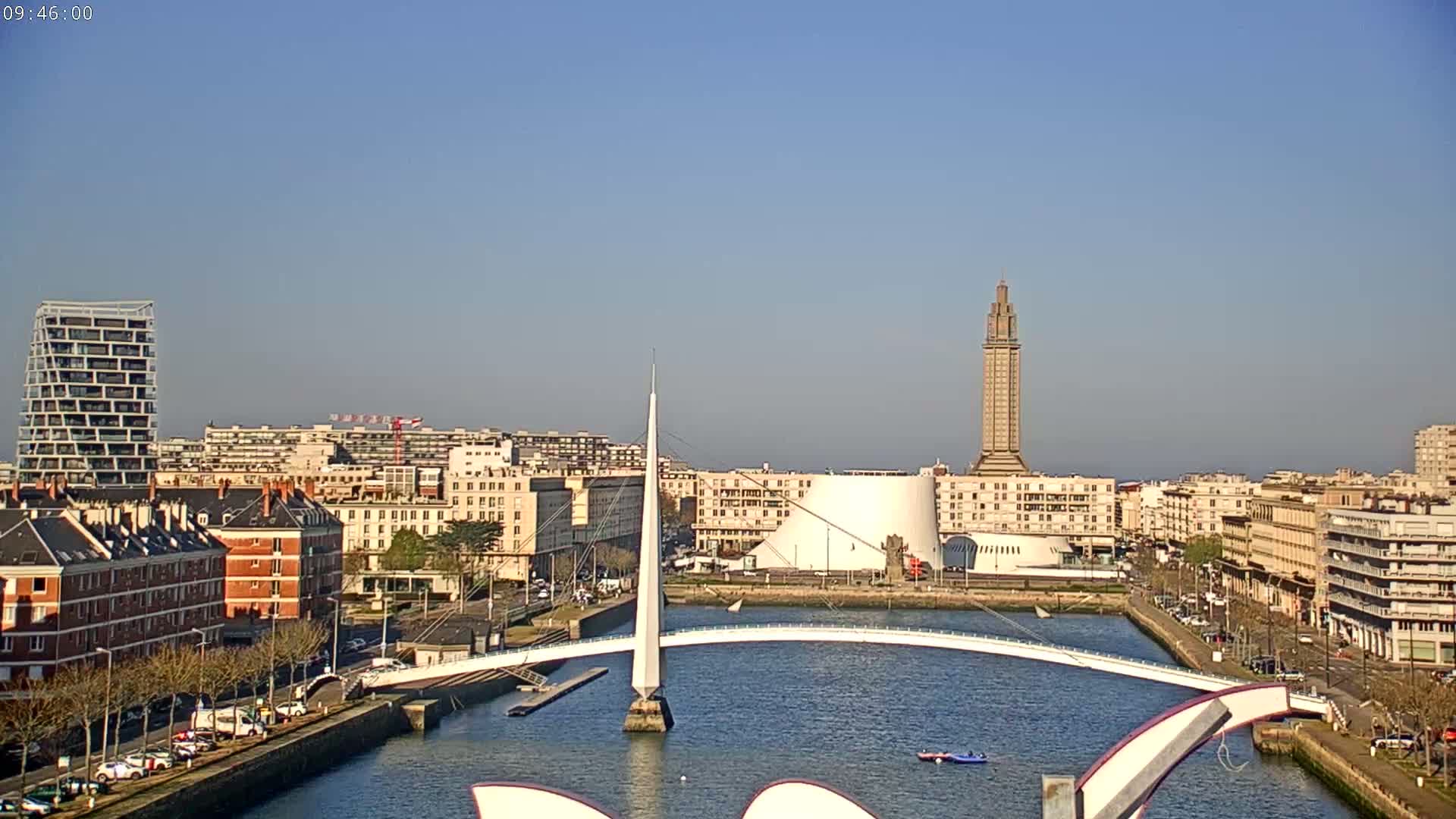 A city waterfront on a sunny day shows a white pedestrian bridge arching over a canal, with buildings and a tall tower in the background.