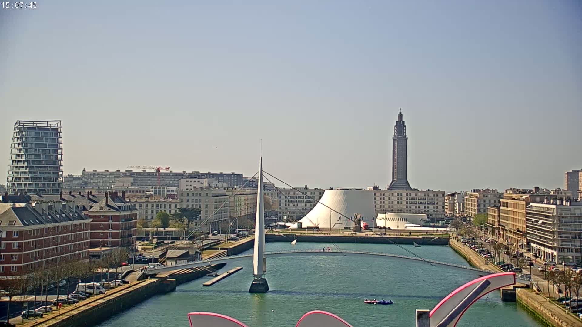 A cityscape with a canal, a modern footbridge, and a tall tower under sunny skies.