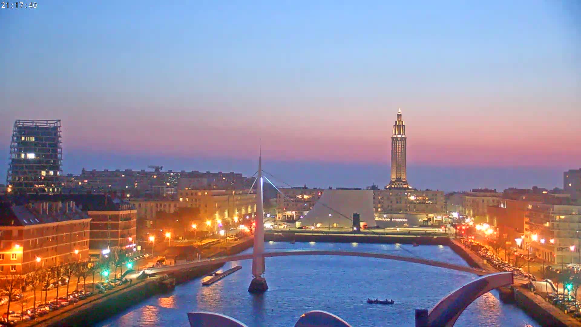 A city at twilight, illuminated by streetlights, with a tall tower in the background and a bridge spanning a canal.