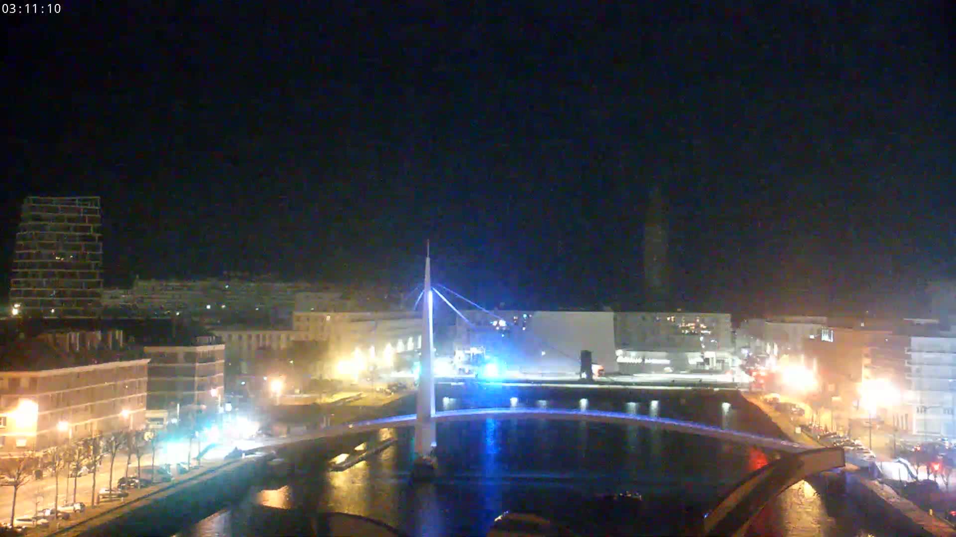 A nighttime view of a city with a lit bridge spanning a canal, under a clear, dark sky.
