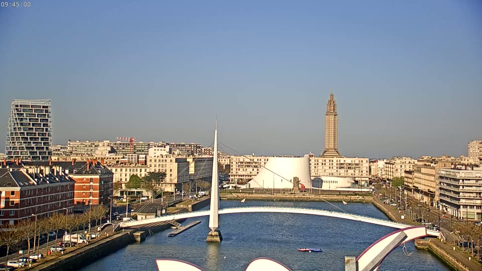 A city waterfront on a clear, sunny day features a modern white pedestrian bridge spanning a waterway, with various buildings, including a tall tower, in the background.