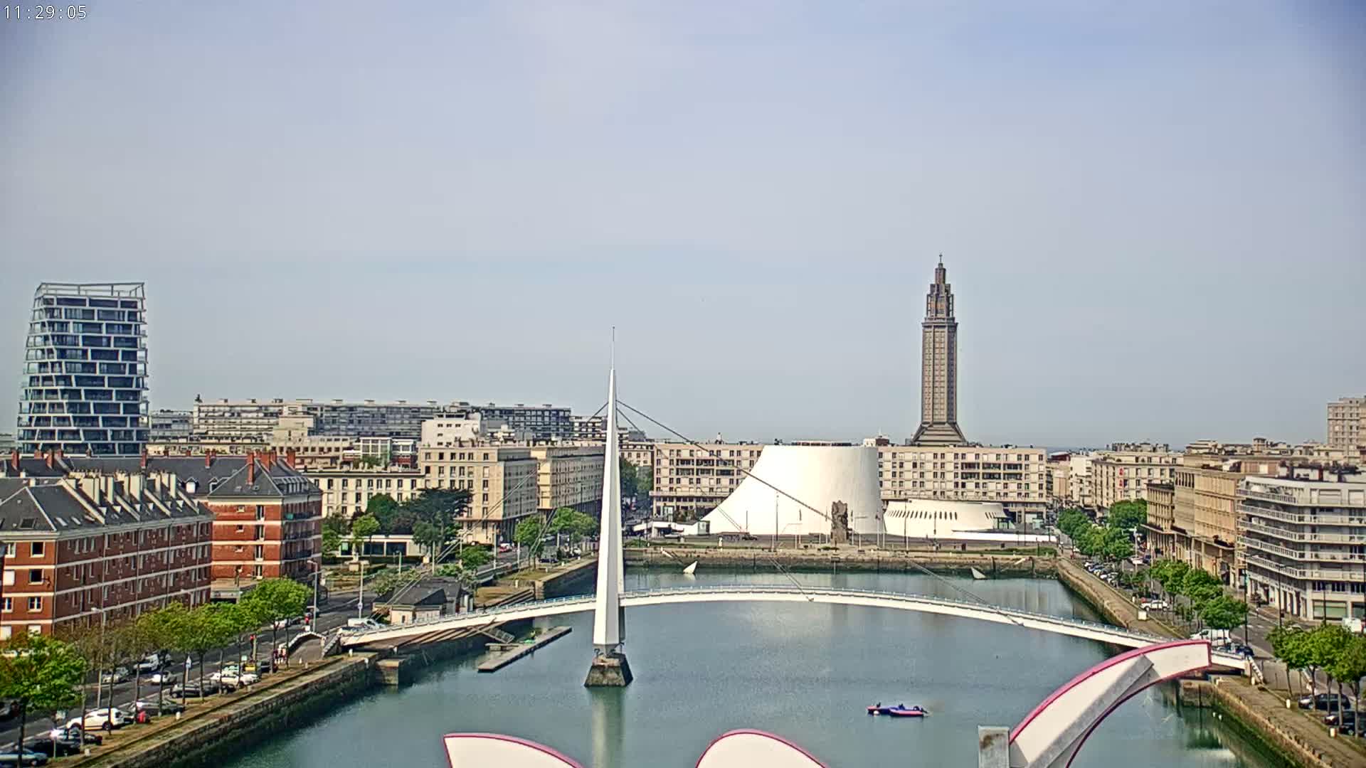 A city skyline on a partly cloudy day features a modern white bridge spanning a canal, various buildings including a tall tower and a dome-shaped structure, and a mix of modern and older architecture.