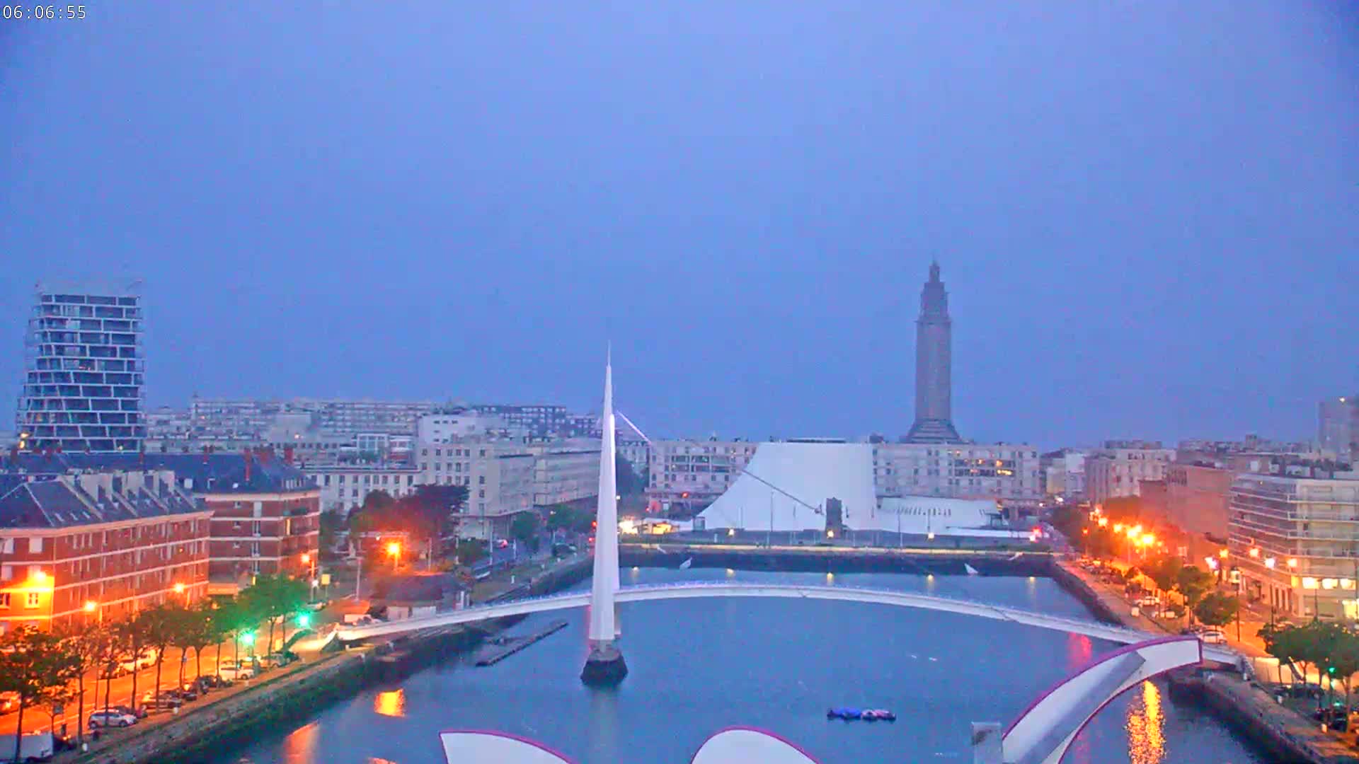 A city at twilight, under a clear, dusky sky, is seen from across a body of water, featuring a modern, white bridge and a tall tower in the distance.
