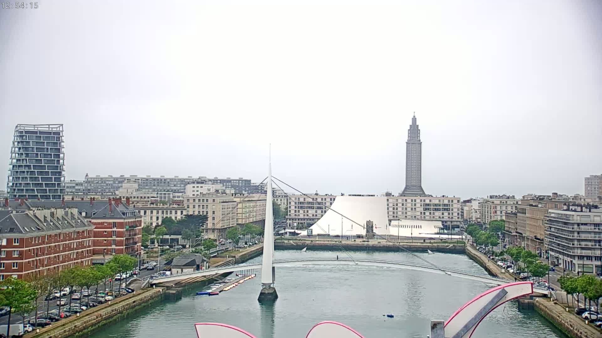 A city waterfront on an overcast day shows a white pedestrian bridge over a calm waterway, with buildings and a tall tower in the background.
