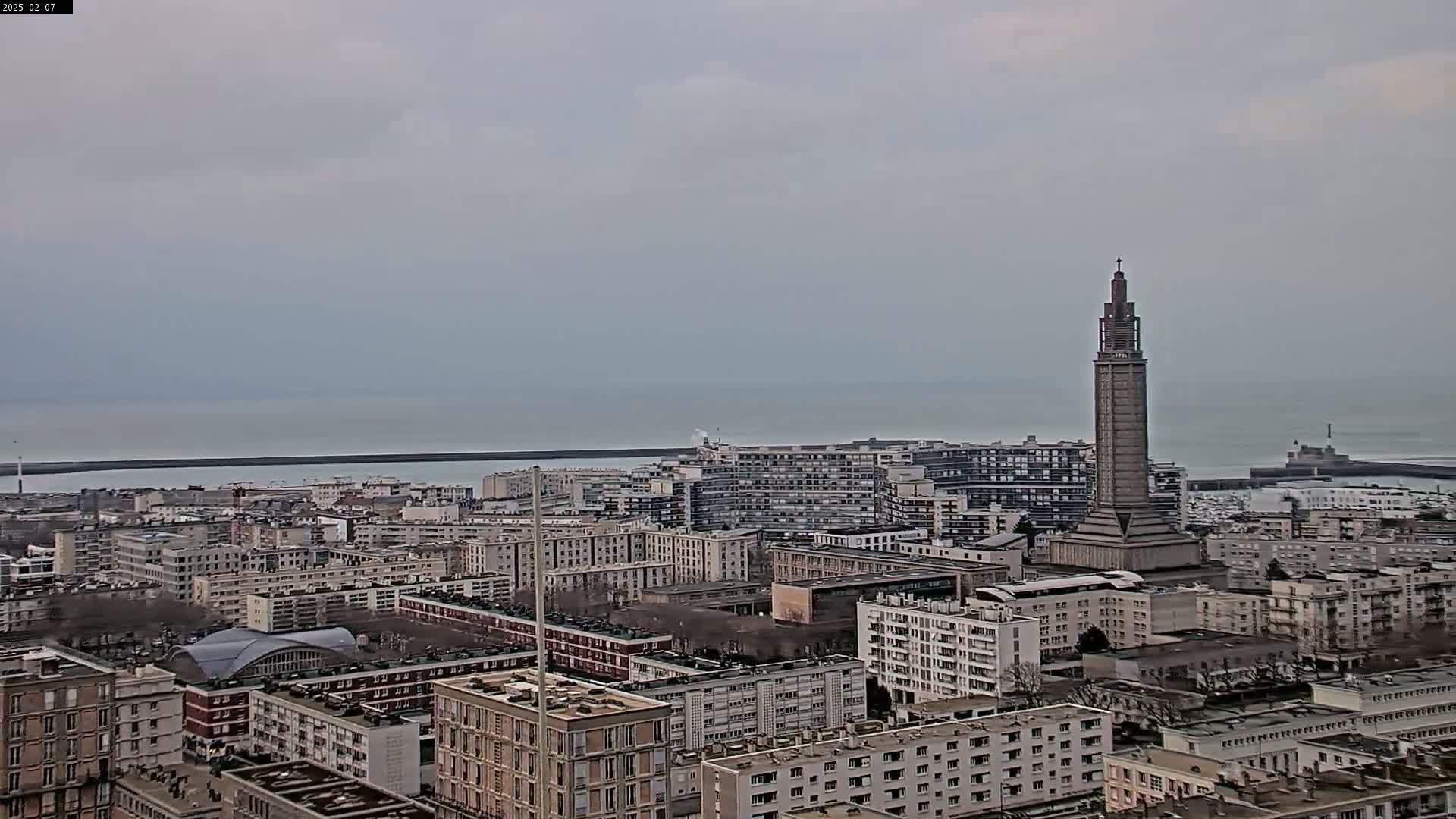 A cityscape with numerous buildings extends to a calm, gray ocean under an overcast sky, featuring a tall, slender tower in the midground.