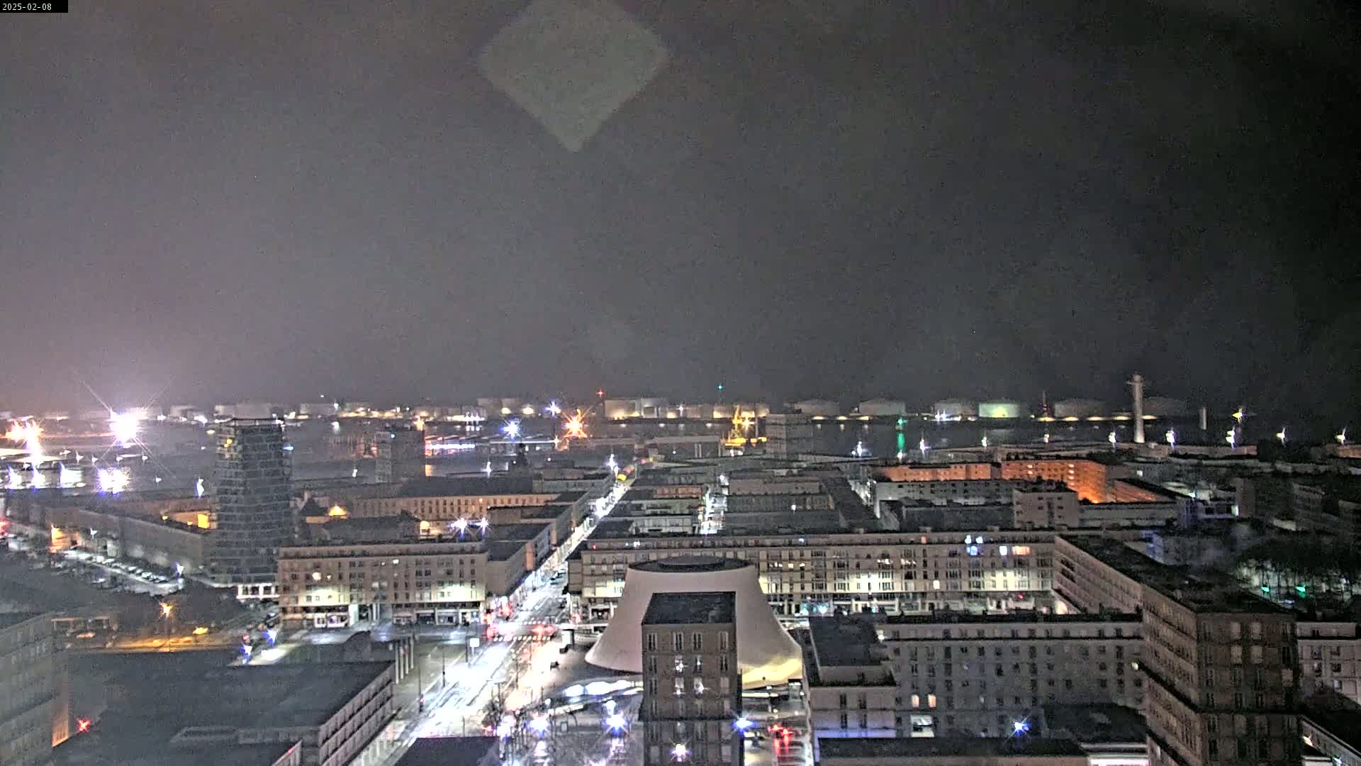 A nighttime aerial view of a city with a harbor in the background under a dark, overcast sky.