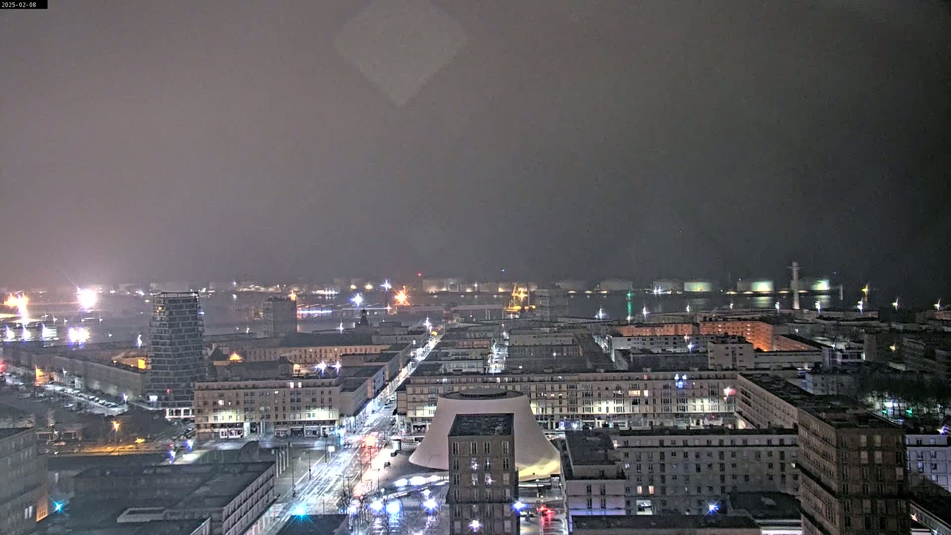 A nighttime, hazy view of a city with a large dome-shaped building near the center, and a harbor visible in the distance.