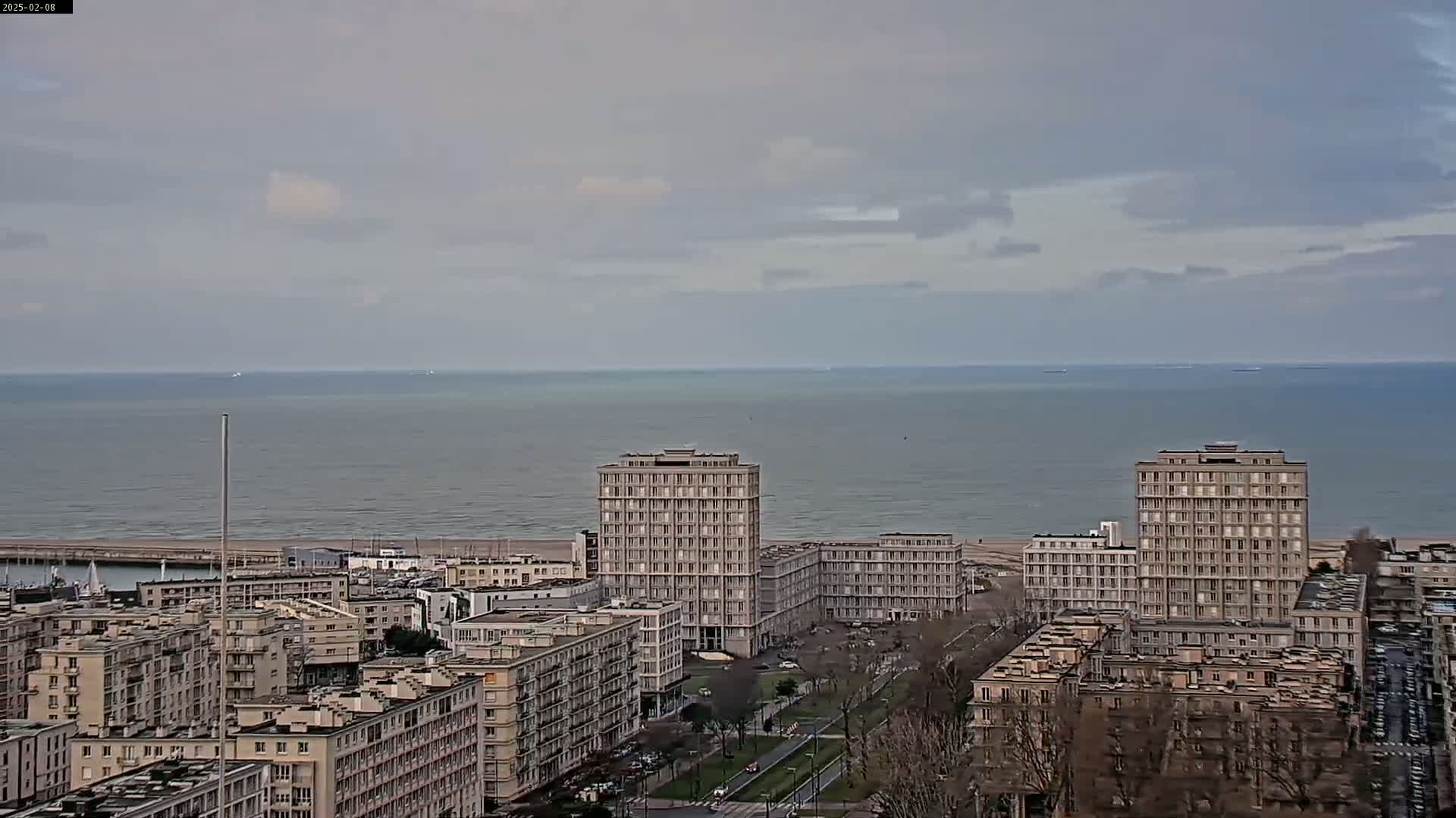 A coastal city with several high-rise buildings is seen under an overcast sky, with a calm ocean in the background.