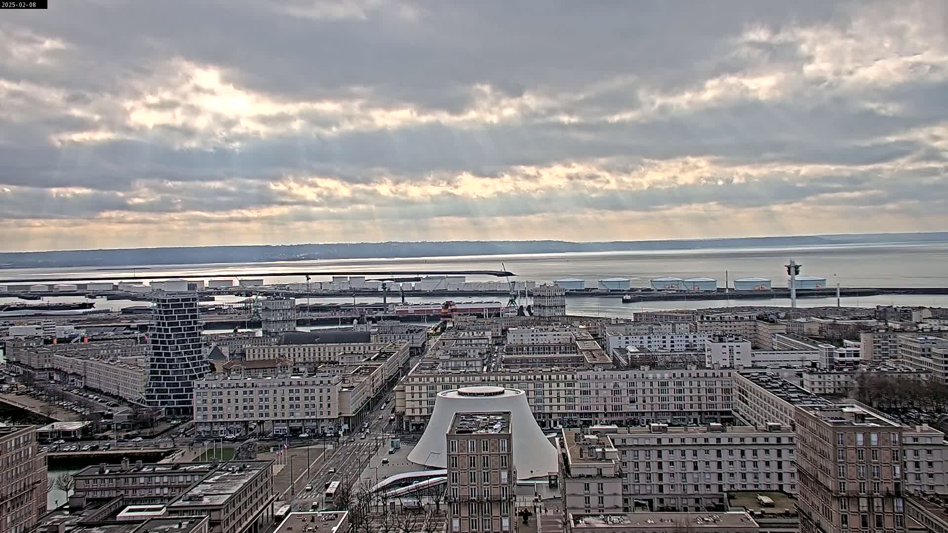A city harbor with numerous buildings and industrial structures is visible under a partly cloudy sky, with sunlight breaking through the clouds.