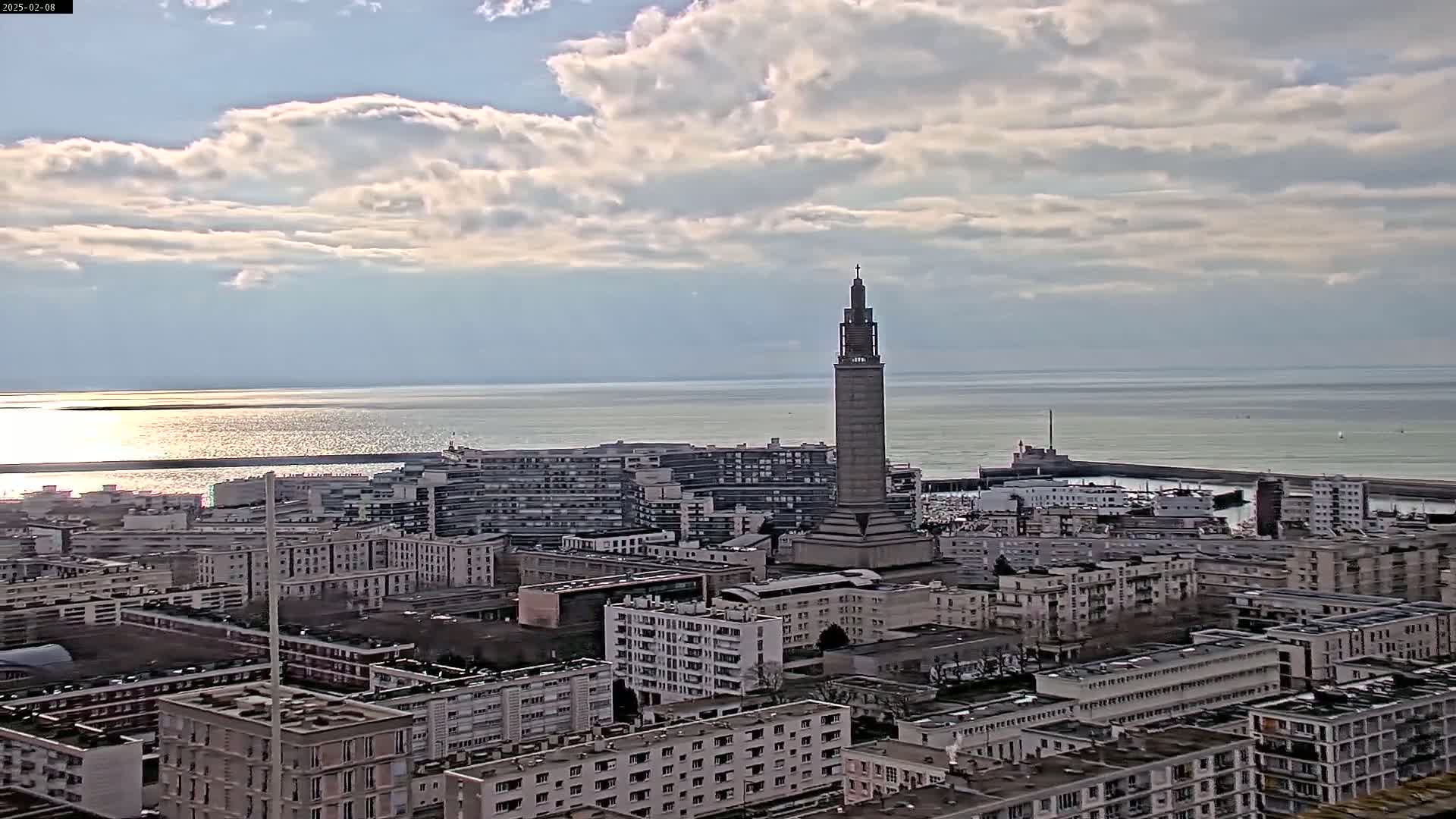 A coastal city with a tall tower is visible under a partly cloudy sky, with calm waters in the background.