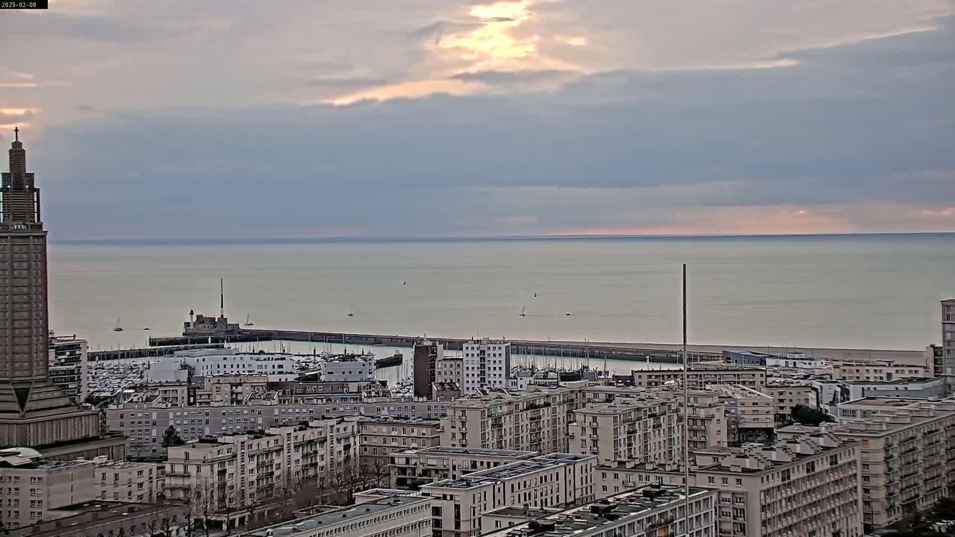 A coastal city with a harbor filled with boats is seen under a cloudy sky with a hint of sunset, as viewed from a high vantage point.