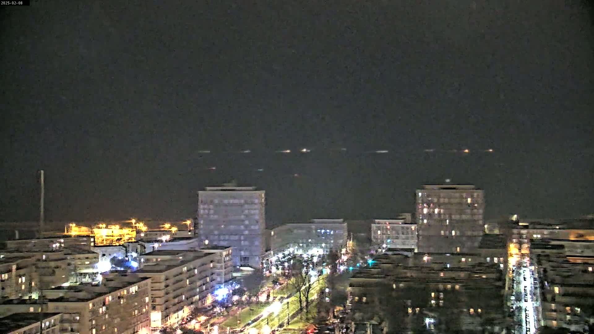 A nighttime view of a city skyline overlooking a dark body of water with several small lights visible in the distance.