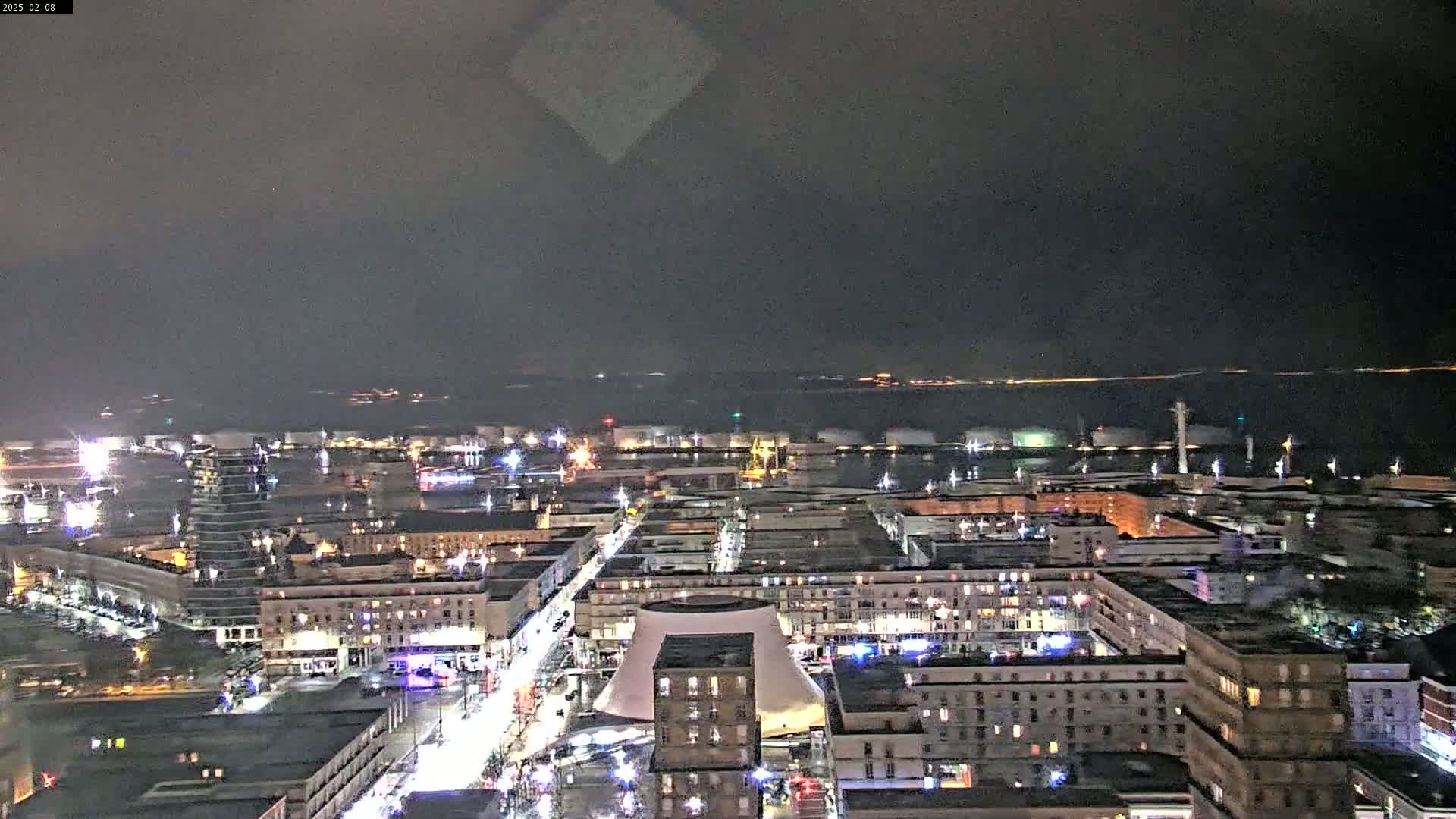 A nighttime aerial view of a city port with illuminated buildings and a body of water in the distance under a dark, cloudy sky.