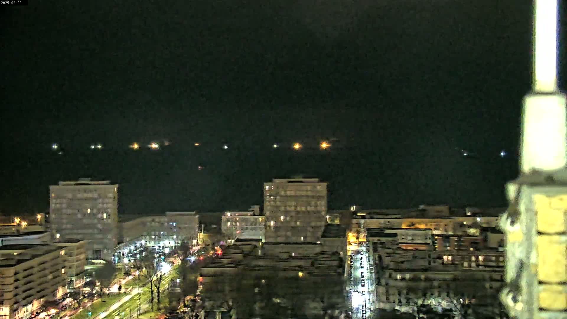 A nighttime, high-angle view shows a city skyline with several multi-story buildings illuminated against a dark sky, featuring a line of distant lights on the water.