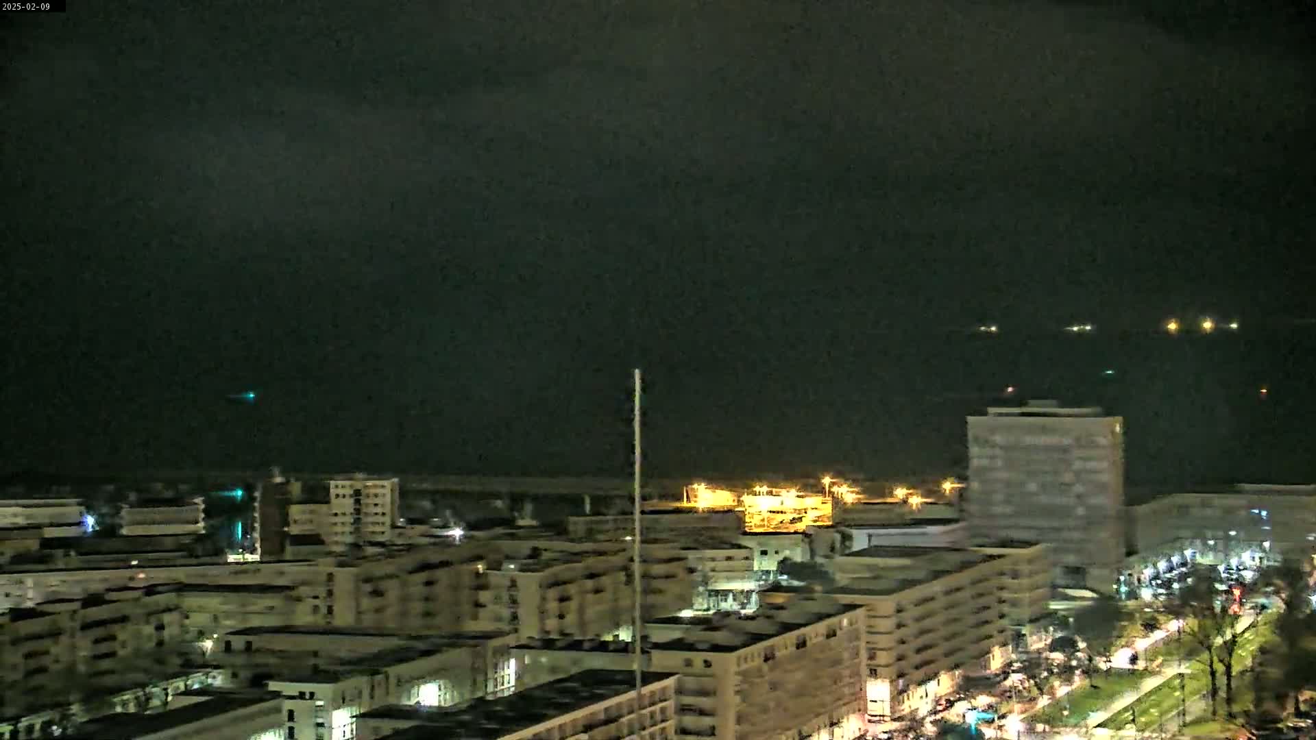 A nighttime, aerial view of a city illuminated by streetlights and buildings, with a dark, cloudy sky and several lights visible on the water in the distance.