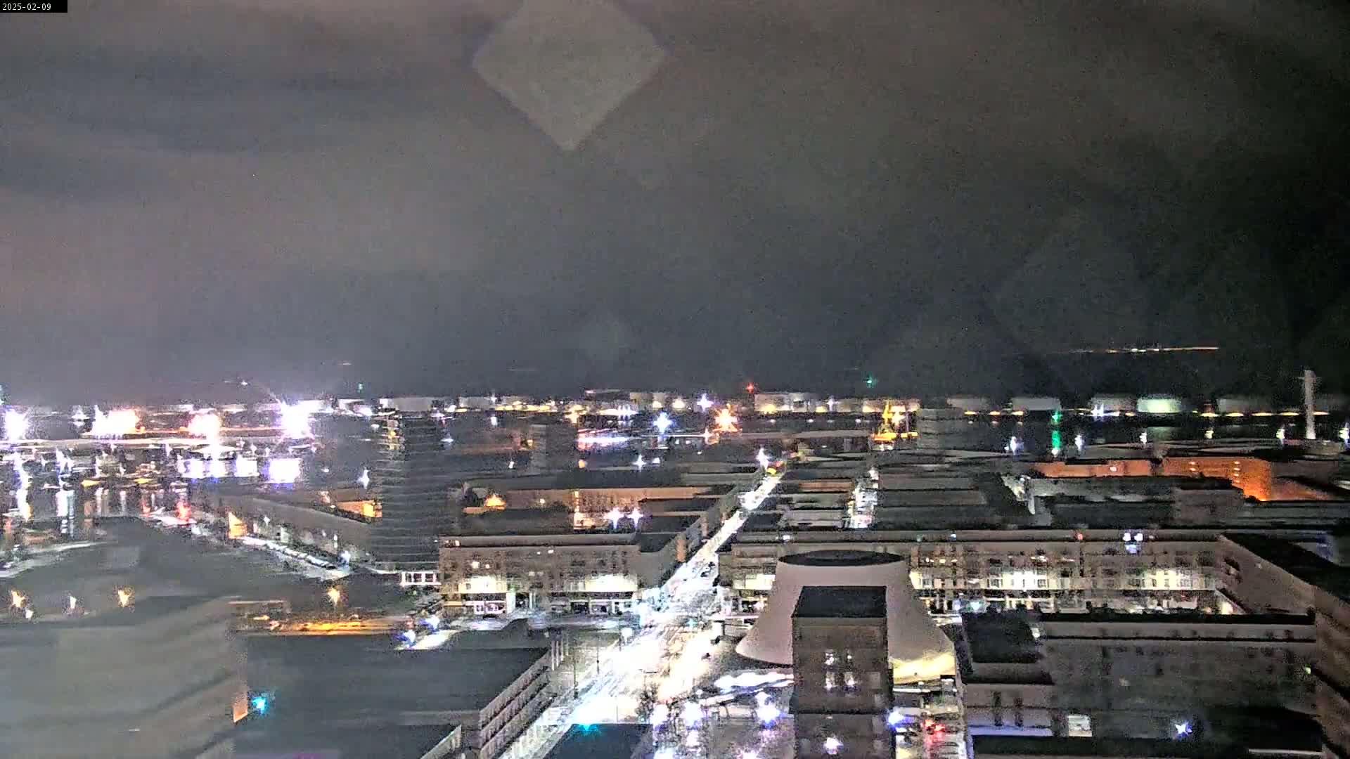 A nighttime, high-angle view of a city with illuminated streets and buildings, under a dark, cloudy sky.
