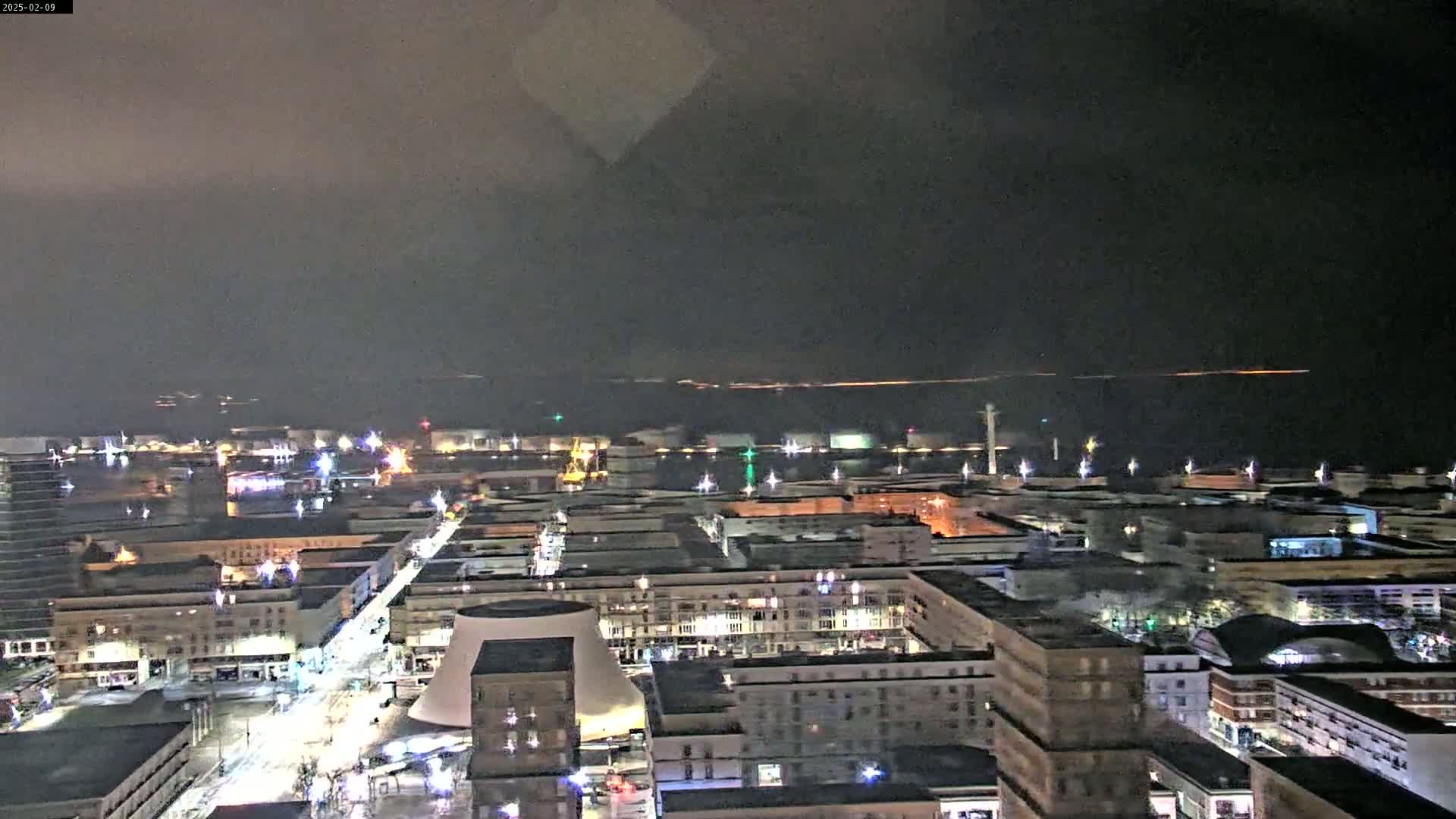 A nighttime aerial view of a city harbor with overcast skies shows a cityscape with lit buildings and streets, and a body of water in the distance.