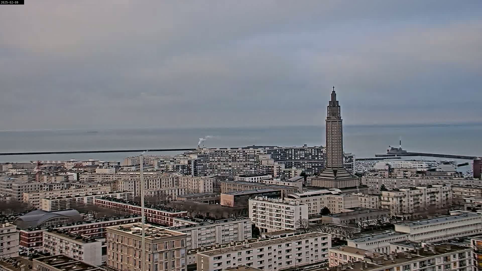 A coastal city with a tall, slender tower is visible under an overcast sky, with a calm sea in the background.