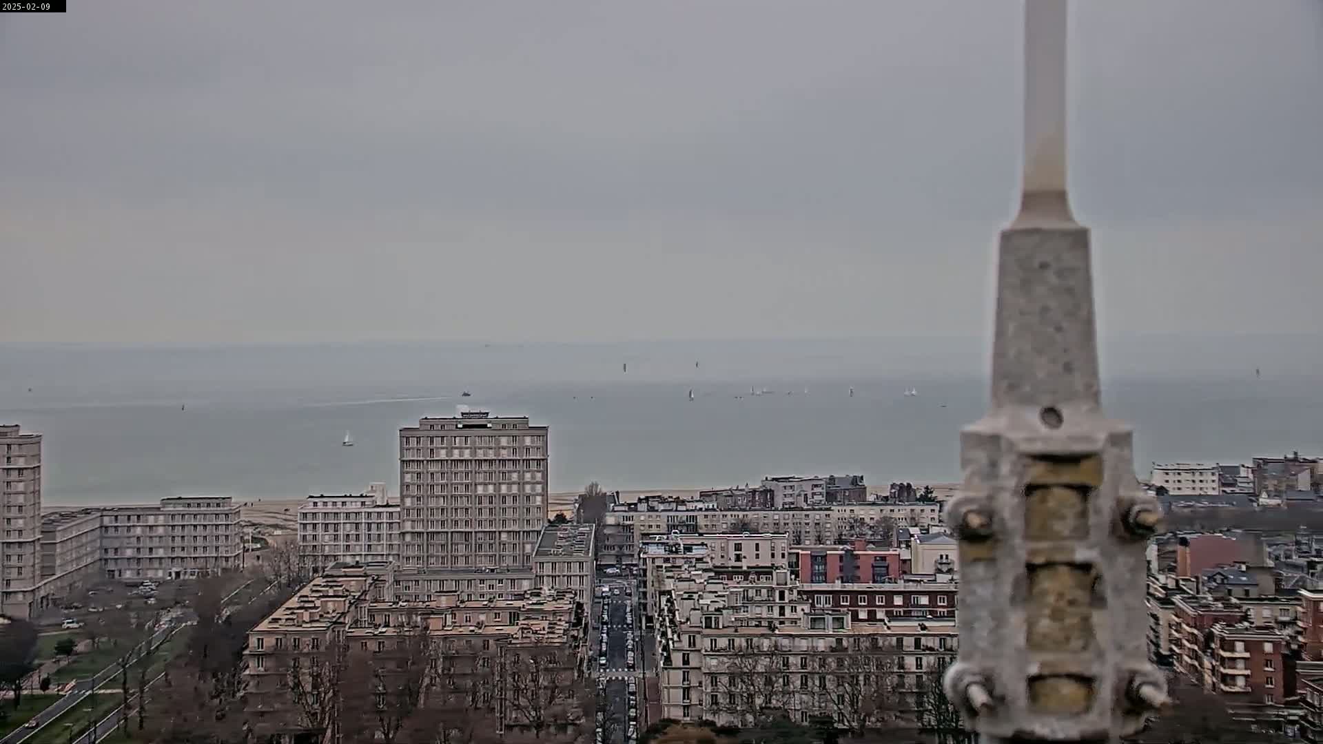 A grey, overcast day reveals a cityscape overlooking a calm body of water dotted with small vessels, with several multi-story buildings prominently featured in the foreground.