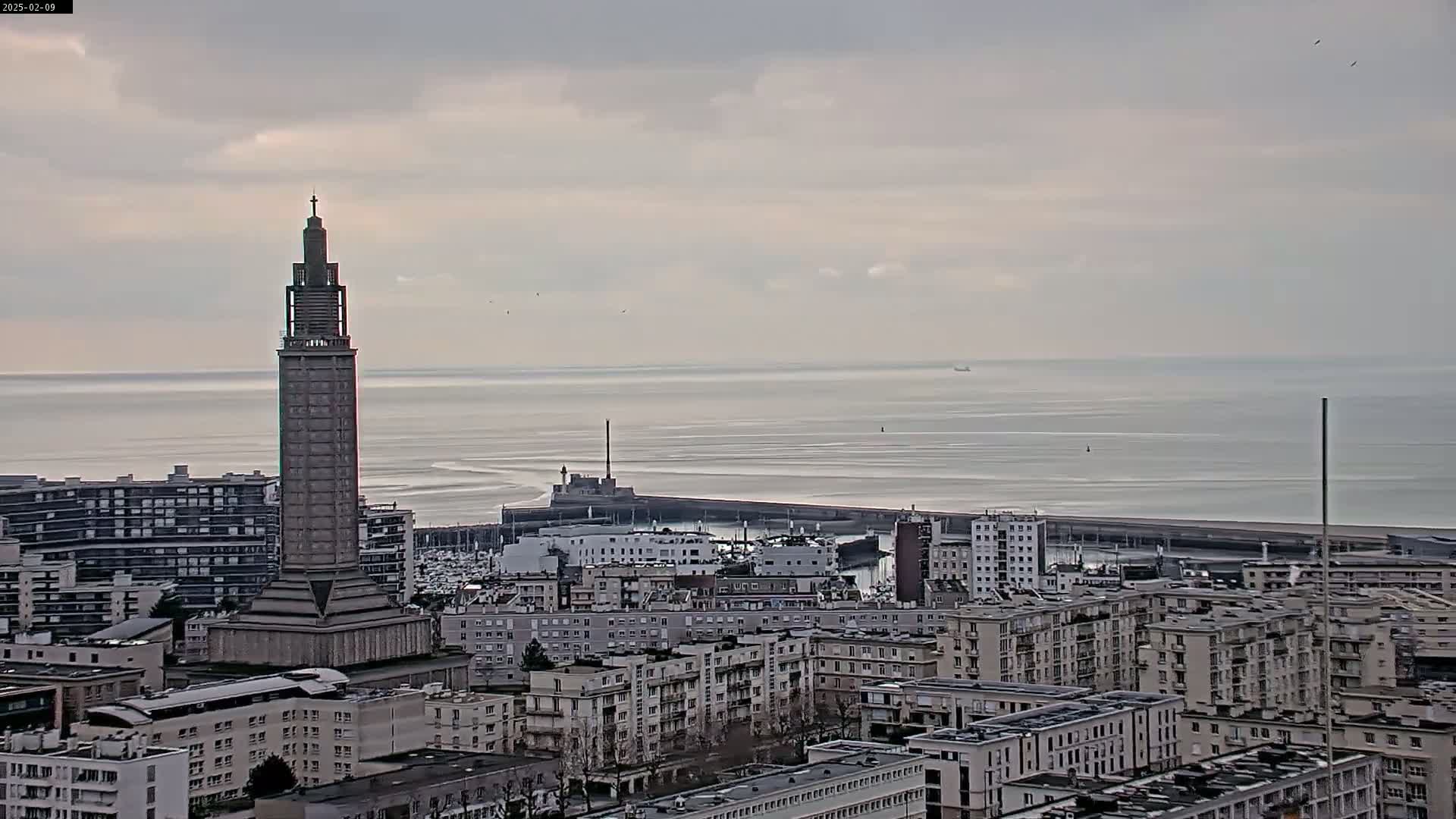 A tall, slender tower stands prominently in a cityscape overlooking a calm, gray sea under a cloudy sky.
