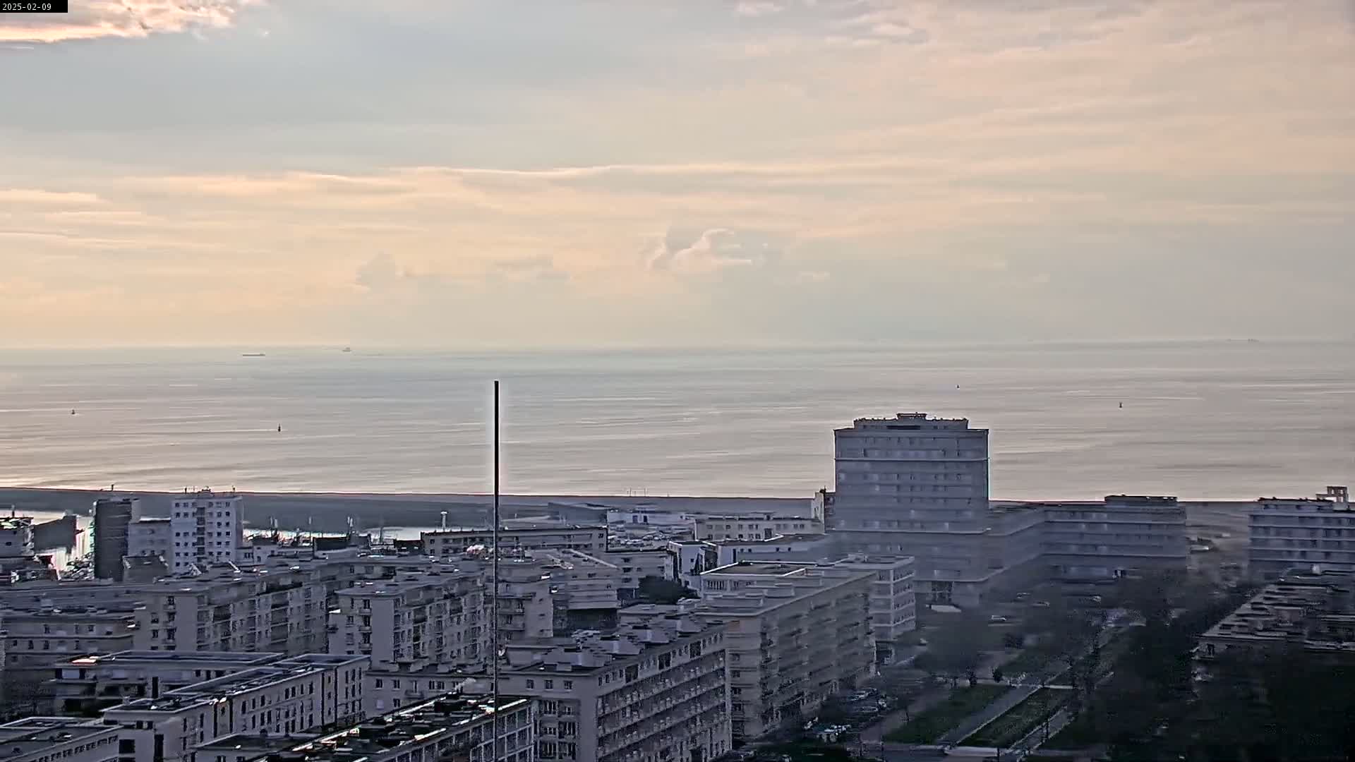 A cityscape of multi-story buildings extends to a calm, gray-blue ocean under a mostly cloudy sky.