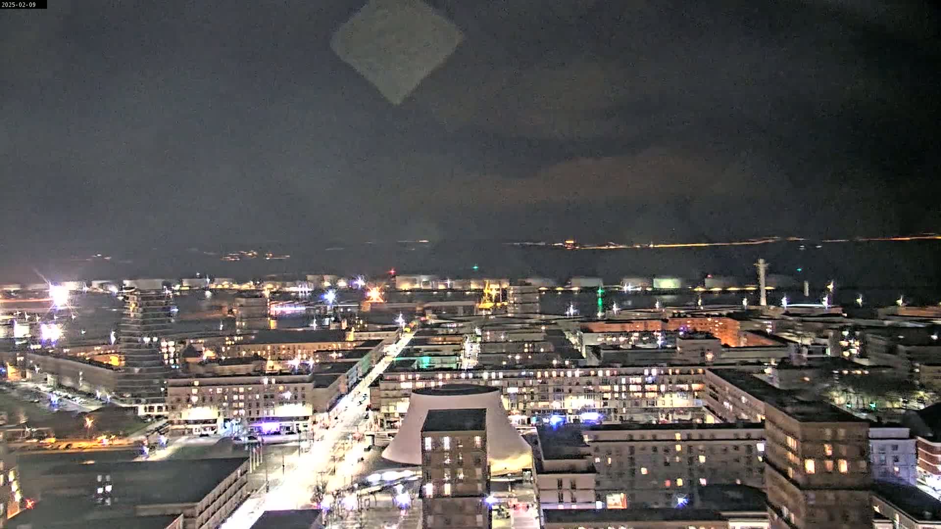 A nighttime aerial view of a city illuminated by streetlights and building lights, overlooking a dark body of water under a mostly clear night sky.