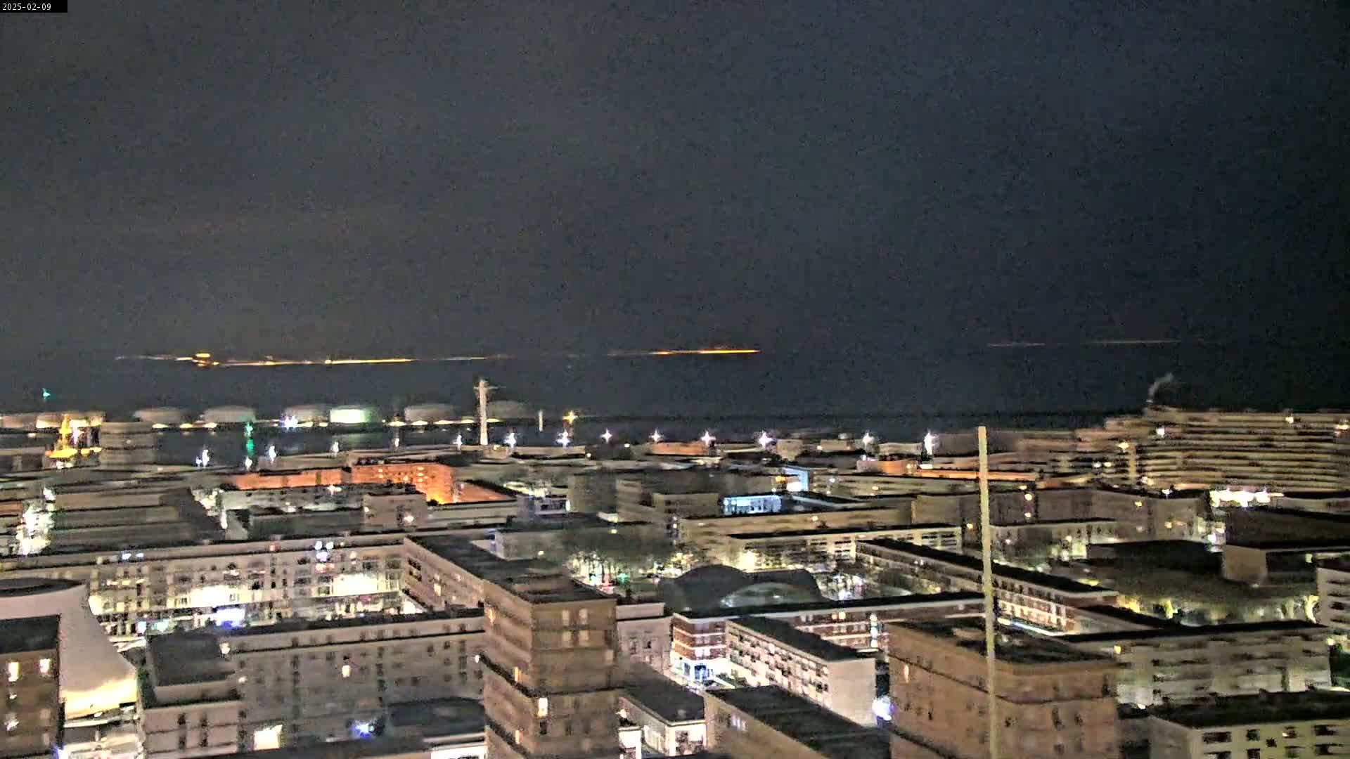 A nighttime view of a city with brightly lit buildings near a dark body of water, showing a faint orange glow on the horizon.