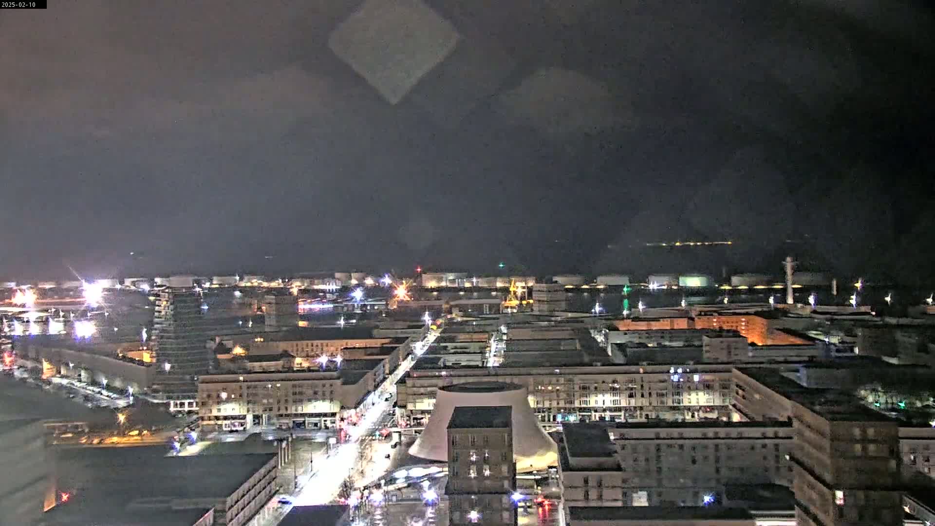 A nighttime cityscape showing city streets and buildings illuminated, with a body of water and industrial structures visible in the distance under a cloudy sky.