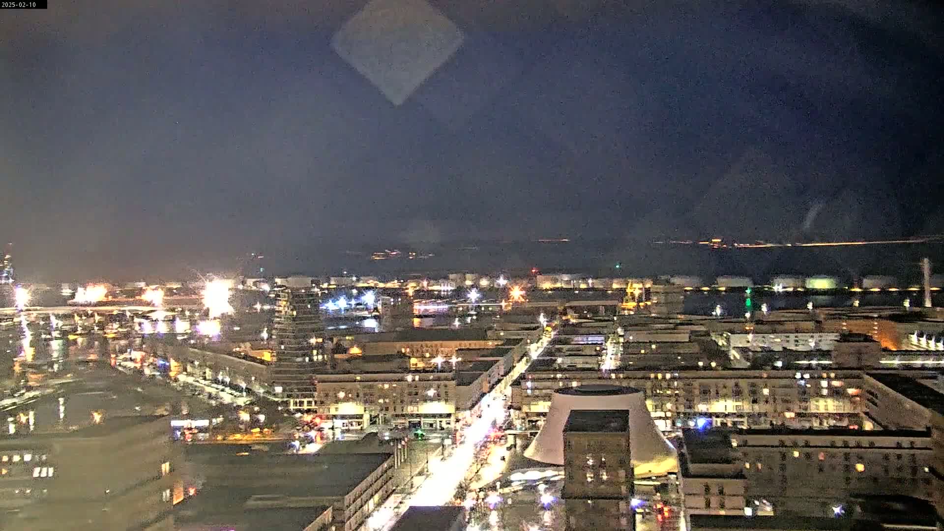 A nighttime, high-angle view of a city harbor with illuminated buildings and streets, under a dark, clear sky.