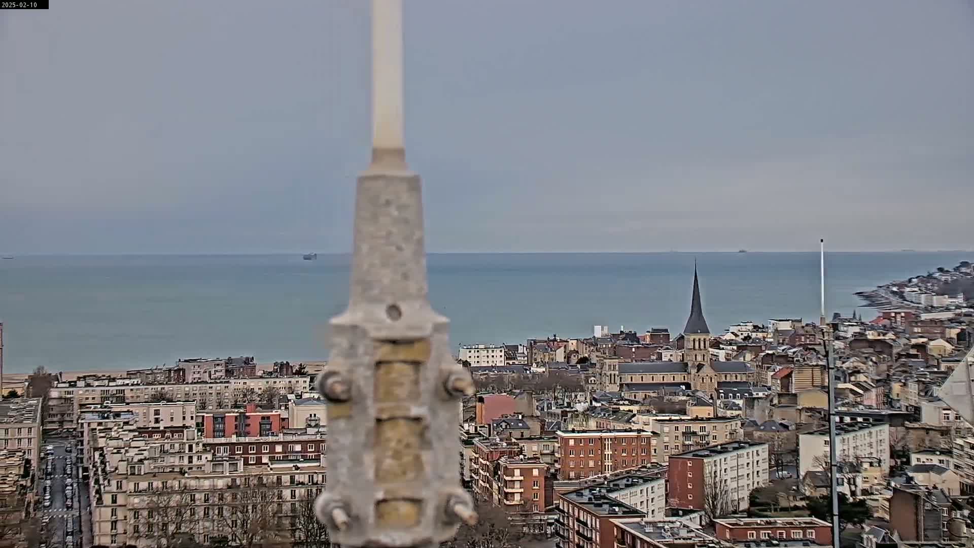 A view from a high vantage point shows a coastal city with a church steeple, calm sea, and overcast skies.