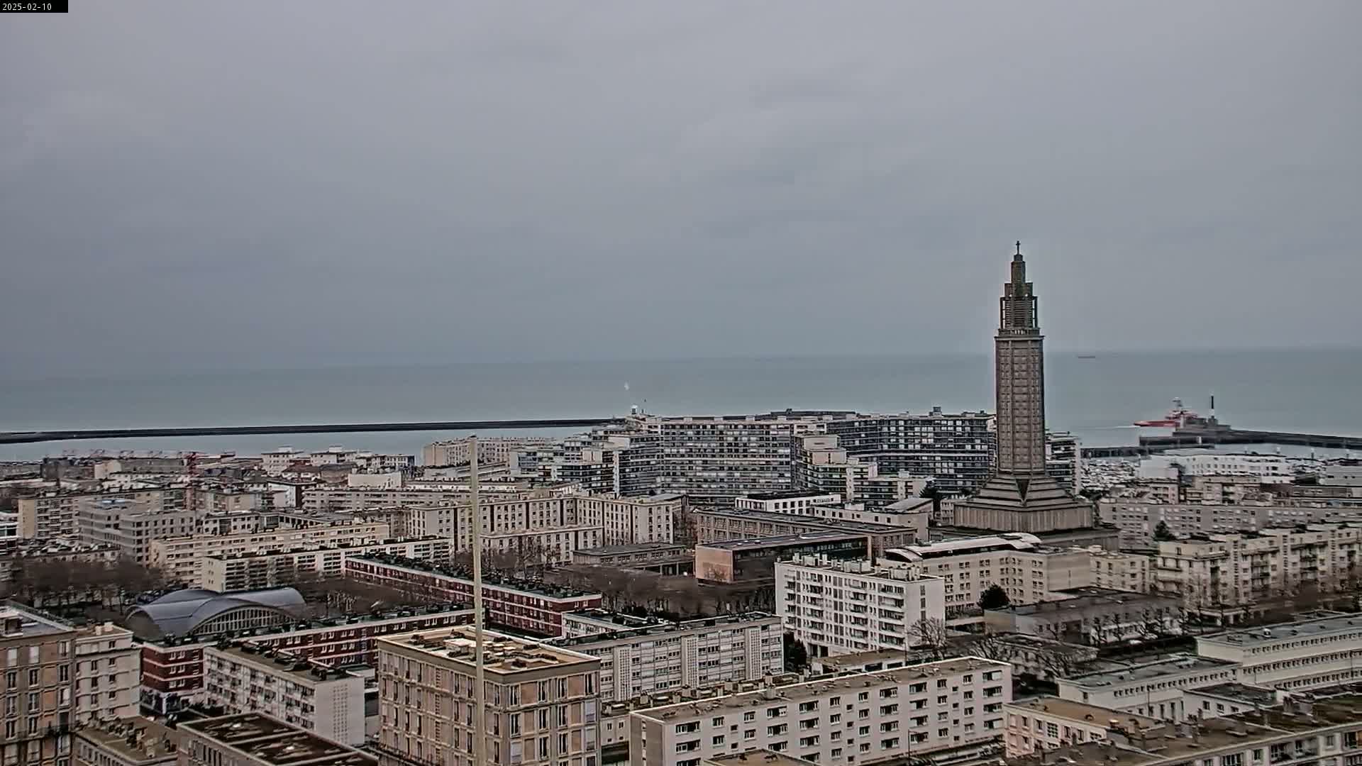 A high-angle, long shot shows a cityscape by the sea on an overcast day, featuring a tall, slender tower among many apartment buildings.