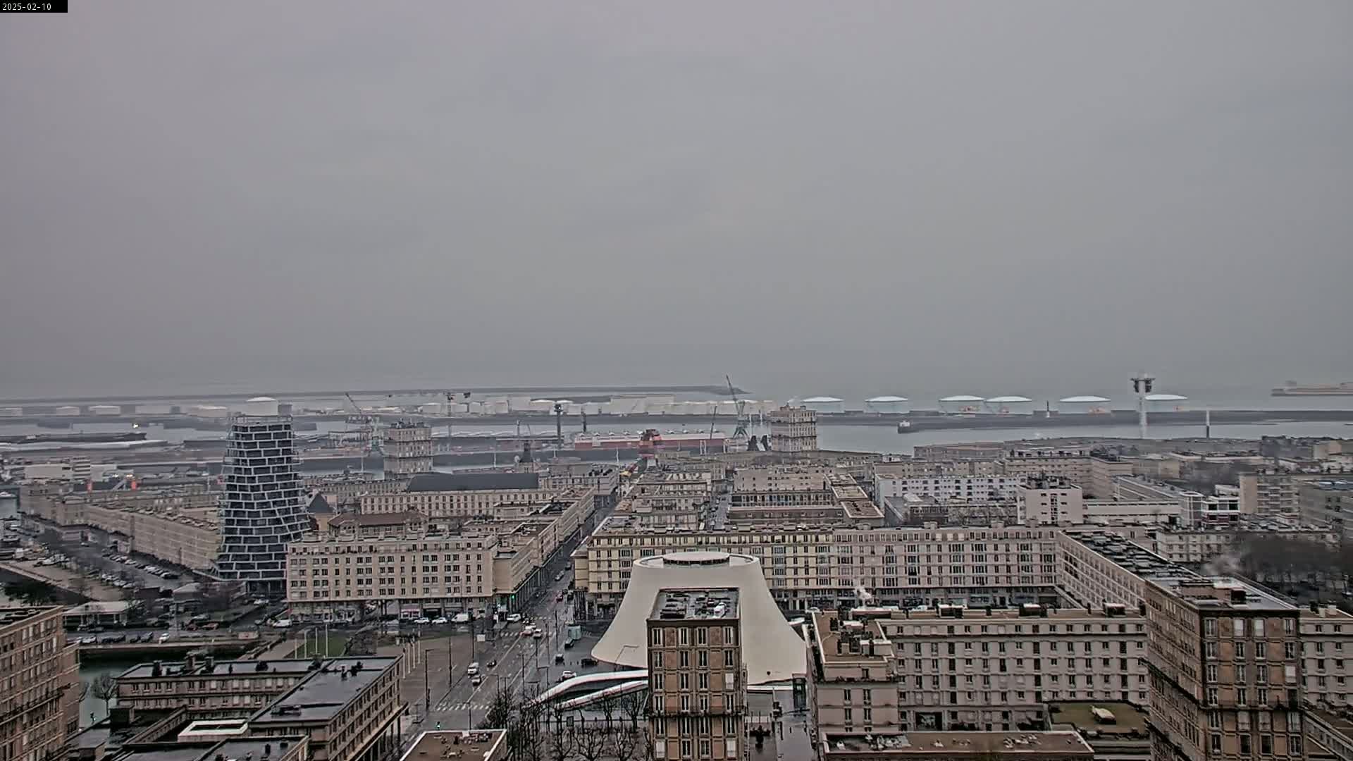 A hazy, overcast view of a city's harbor and cityscape, featuring a prominent modern building and numerous multi-story structures.