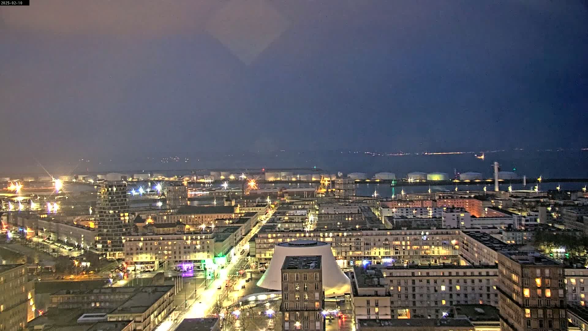 A nighttime aerial view of a city illuminated by streetlights and building lights, overlooking a harbor with industrial structures under a dark, clear sky.