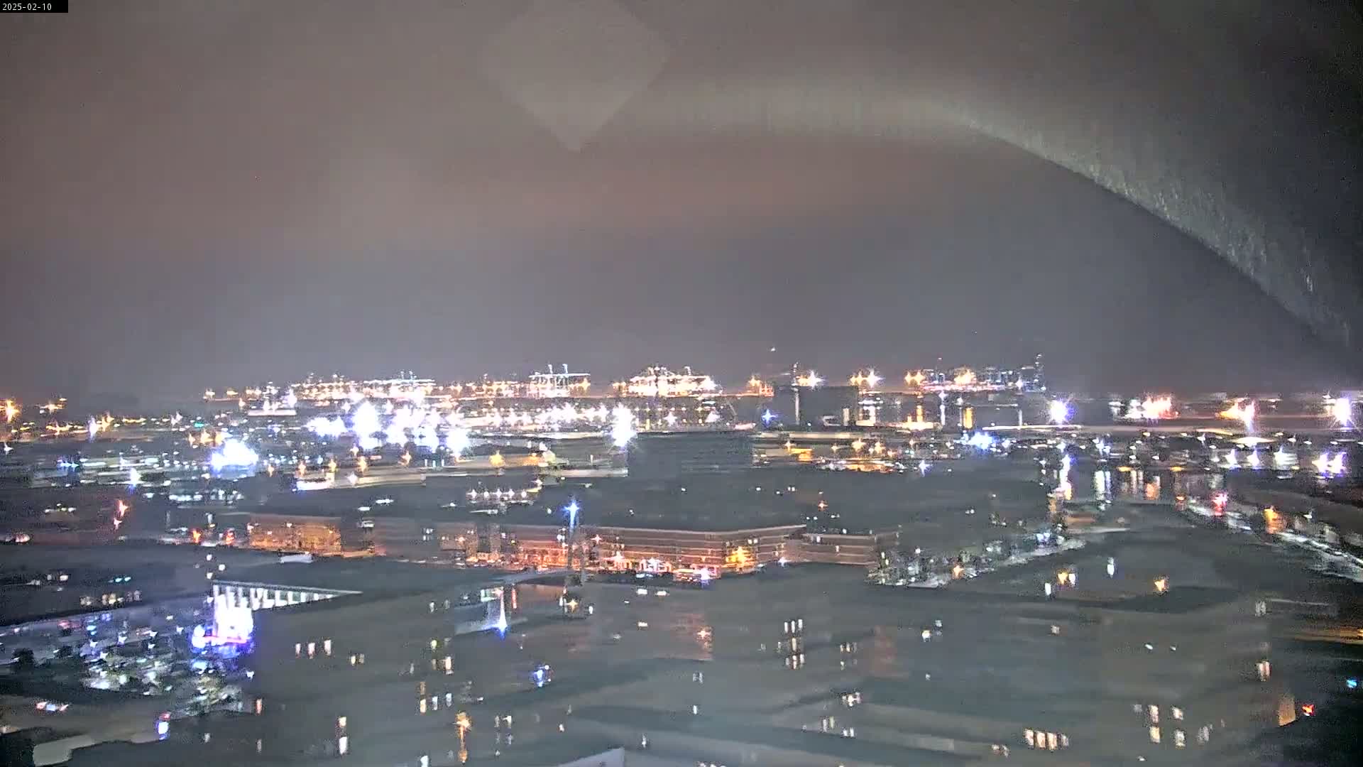 A nighttime view of a city's waterfront, illuminated by numerous lights, under a dark, possibly rainy sky.