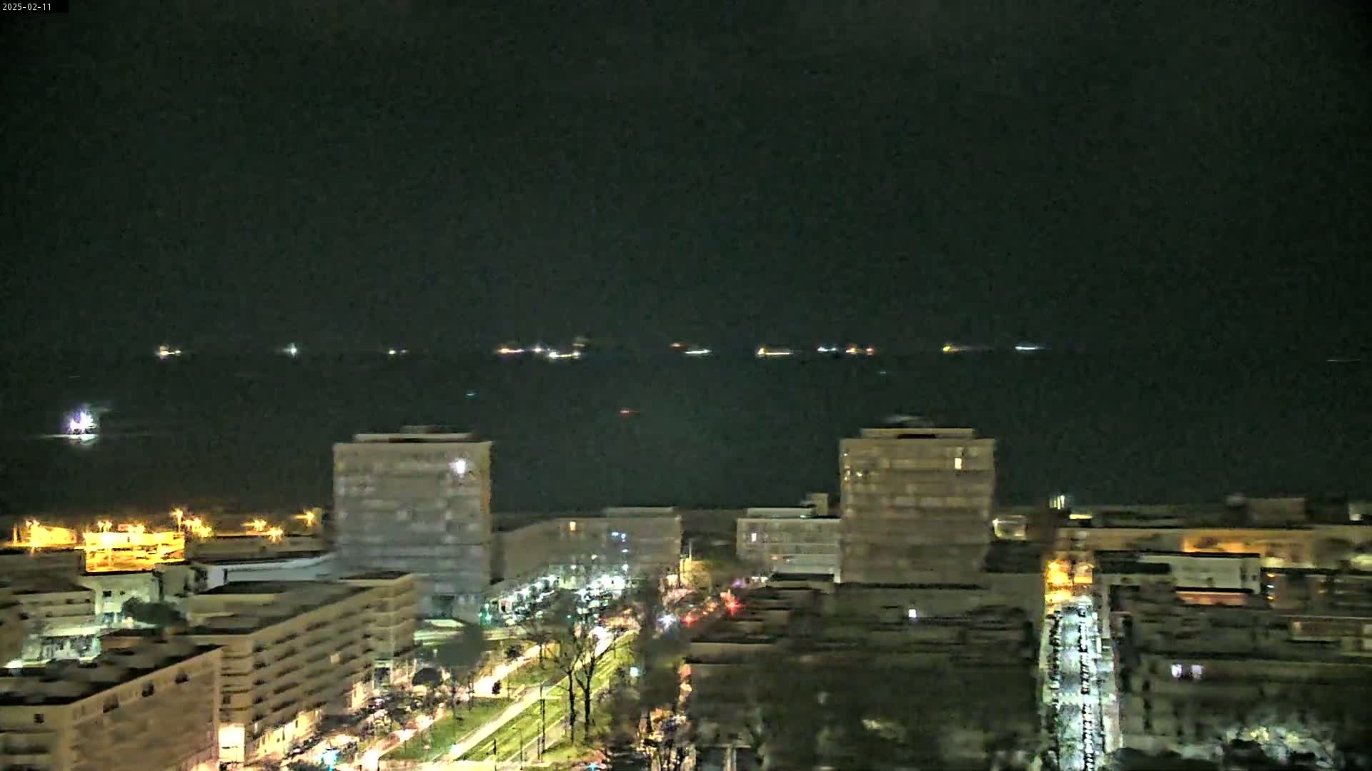 A nighttime, aerial view of a city with several buildings illuminated, overlooking a dark body of water where distant lights suggest ships at sea.