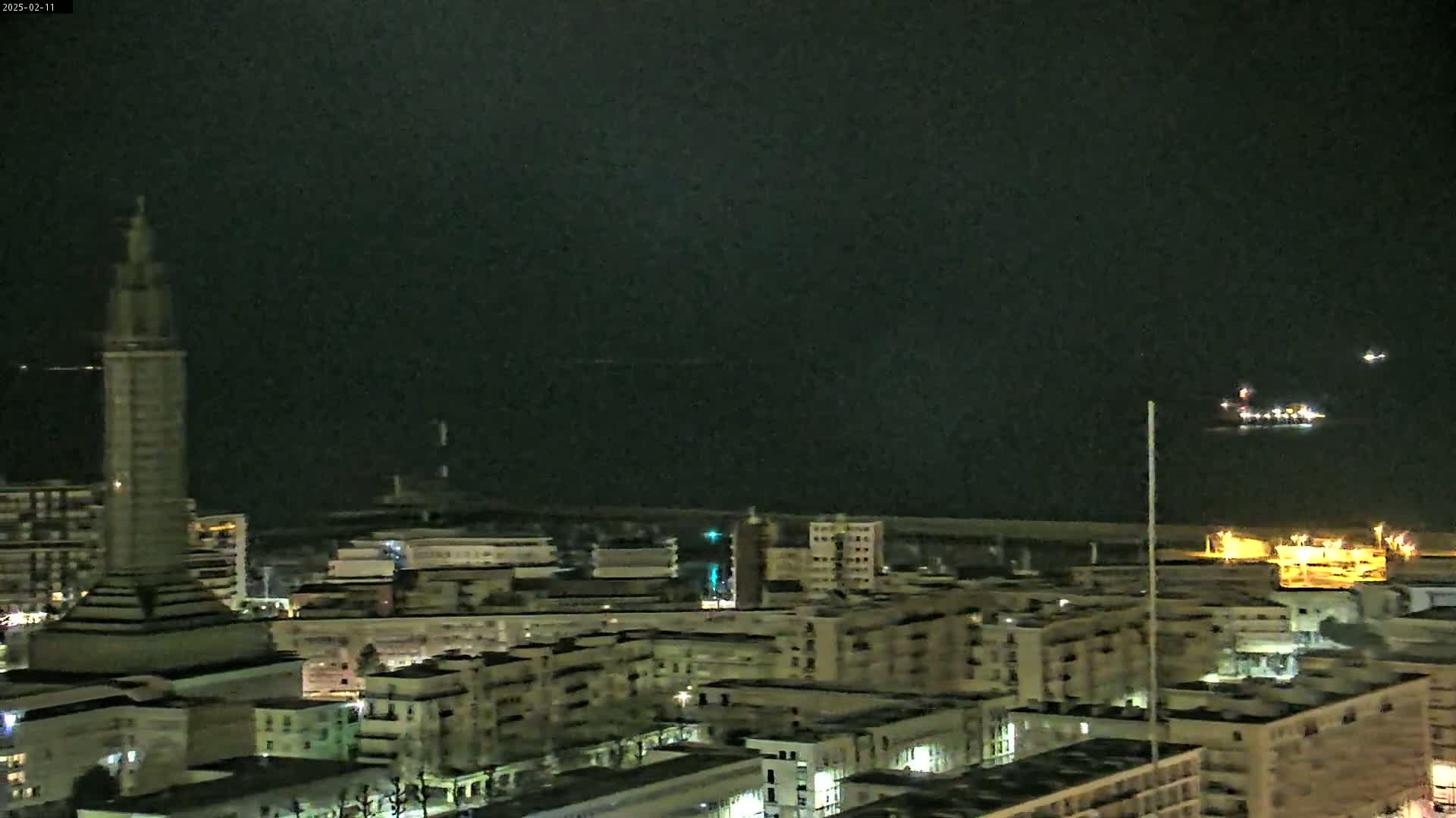 A nighttime view of a city with a tall tower, buildings, and a dark body of water with lights visible in the distance under a dark, clear sky.
